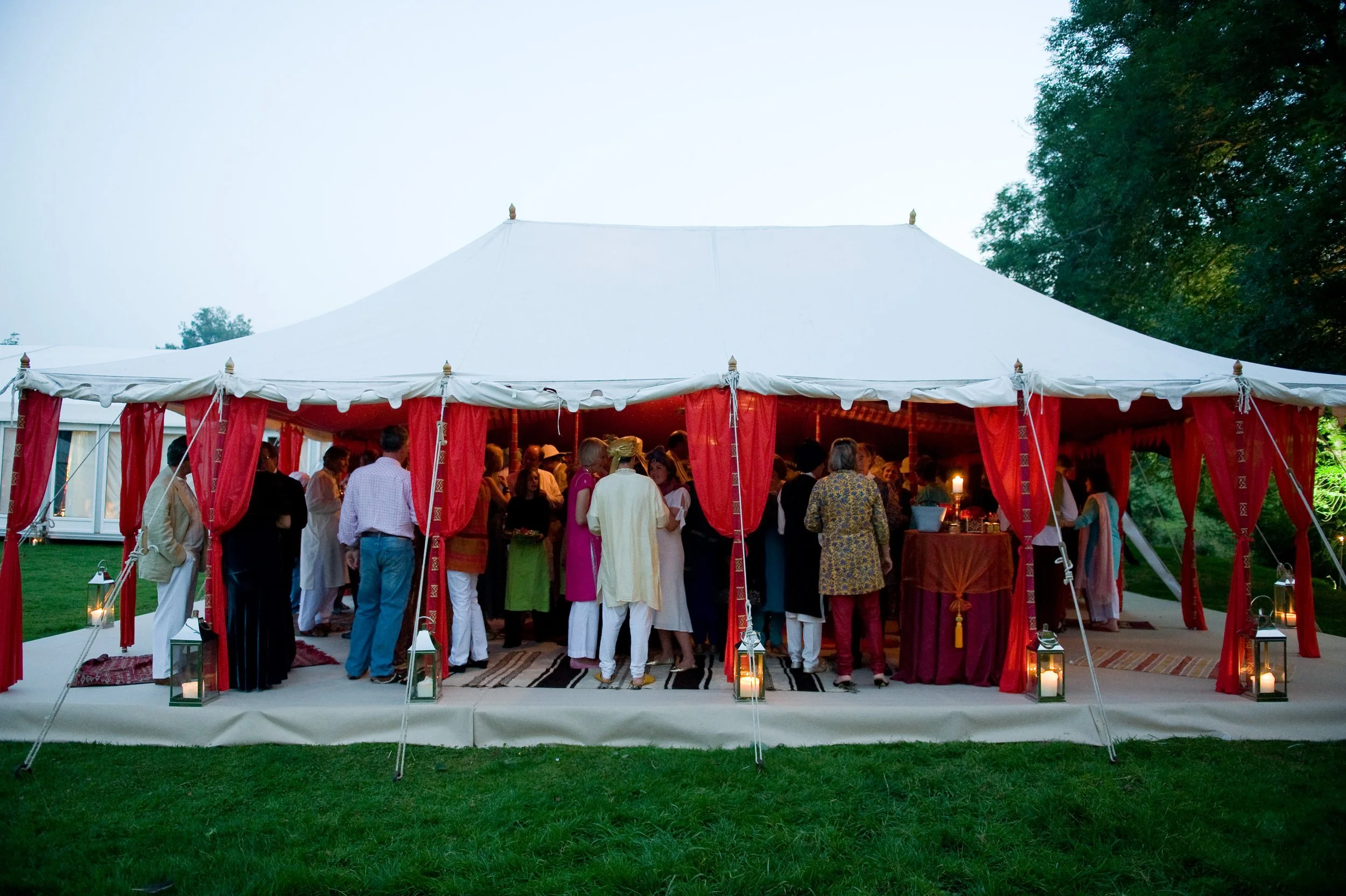 People gathered under a white and red wedding tent at an outdoor event, with lanterns on the grass and trees surrounding the area.