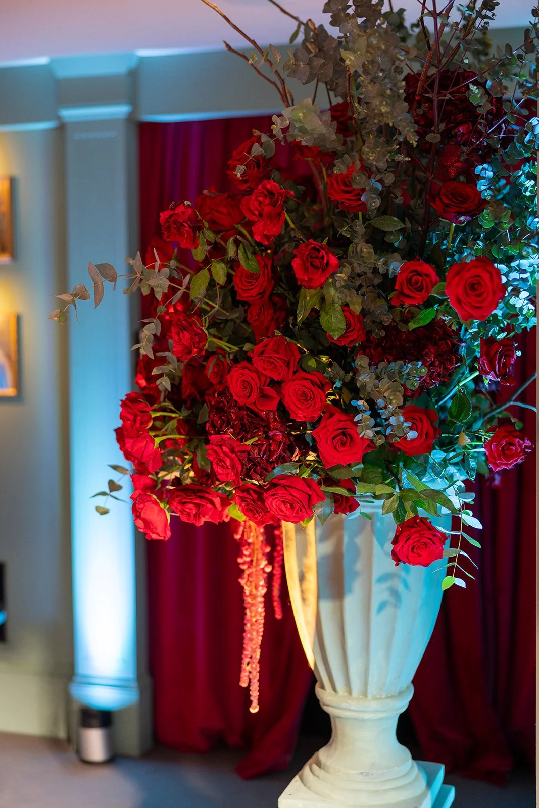 A large vase filled with red roses, green foliage, and other flowers, placed indoors with red curtains and decorative lighting.