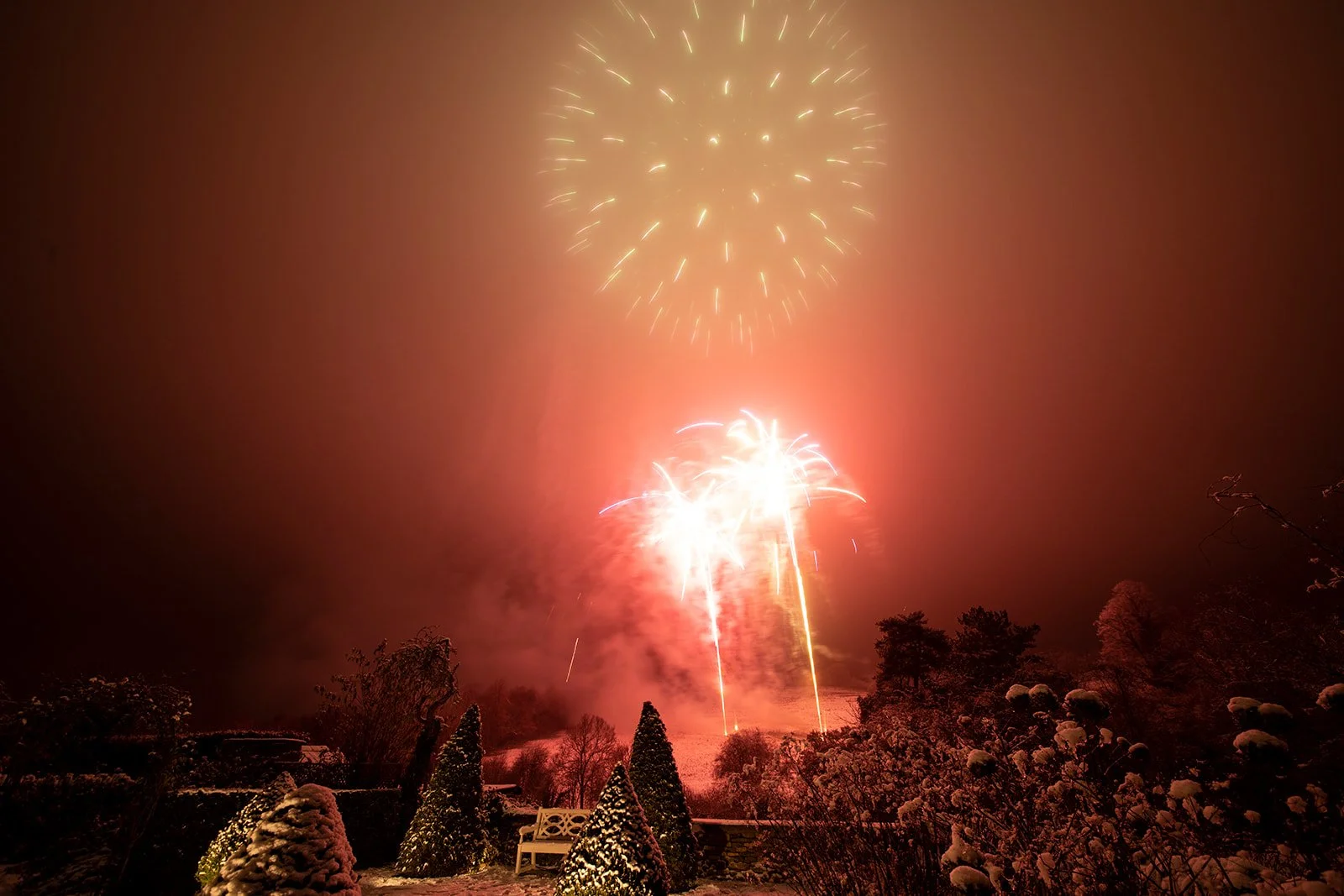 Fireworks lighting up the night sky over a snowy landscape with trees and bushes.