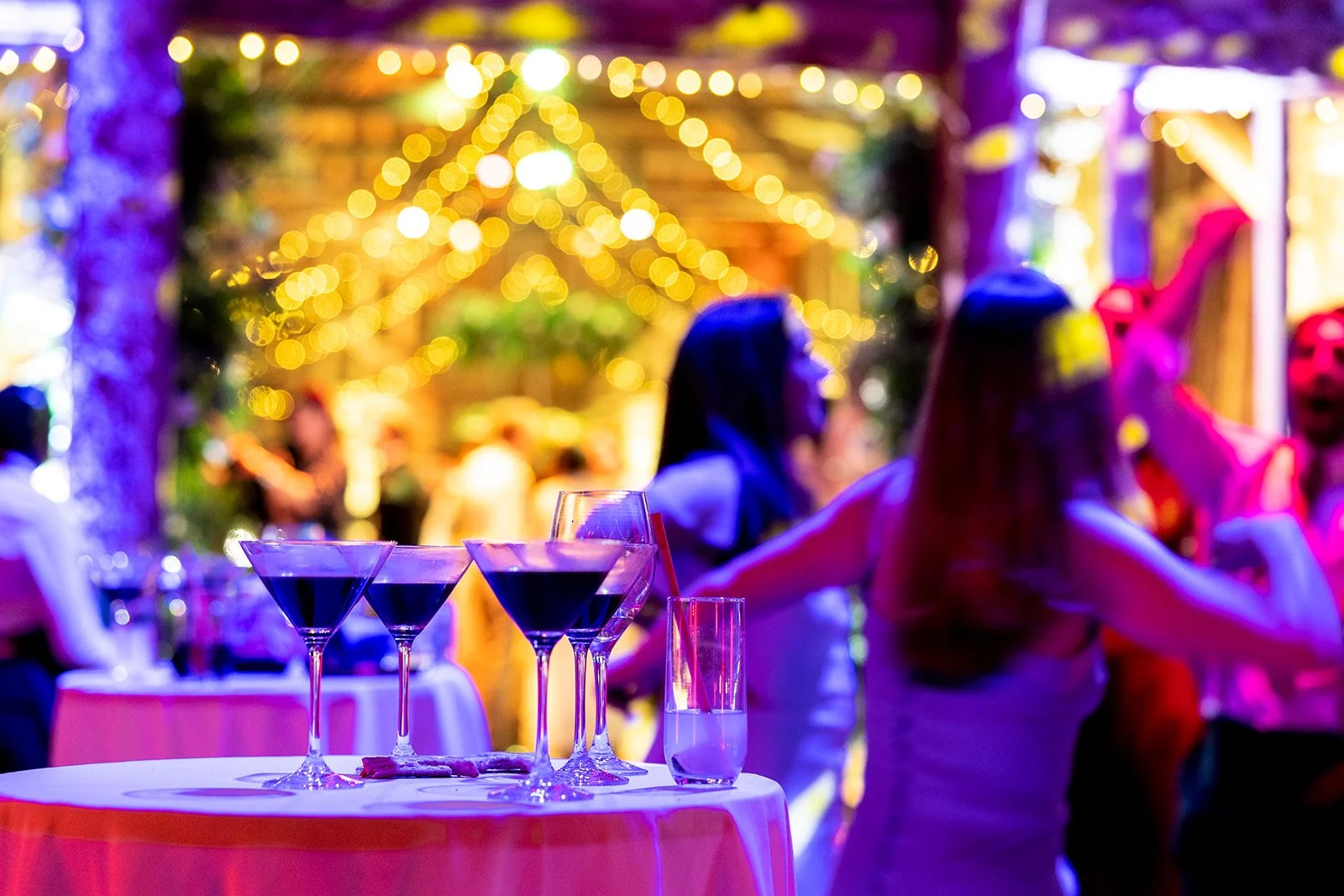 Celebration scene with a table of cocktails and guests dancing under colorful string lights at a festive party.
