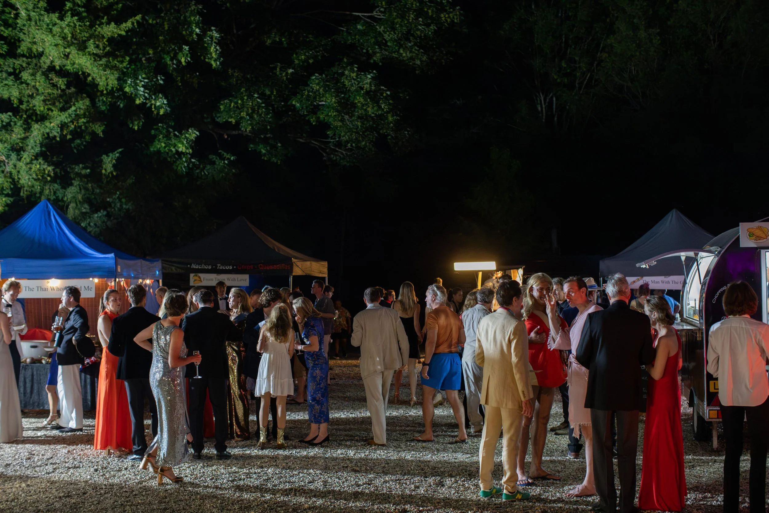 People gathered at an outdoor night event with food stalls and tents, surrounded by trees.