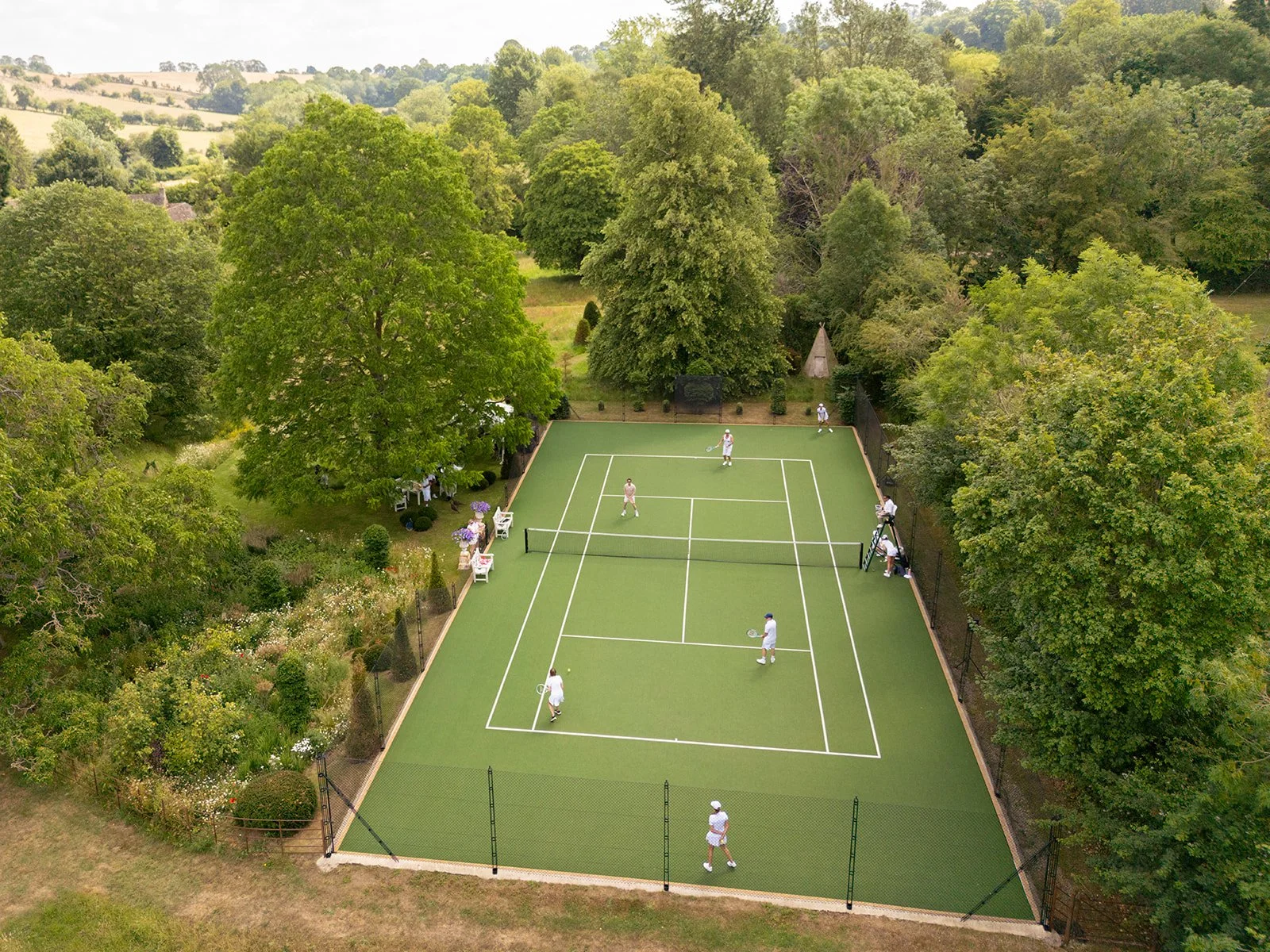 Aerial view of a green tennis court with six people playing, surrounded by lush trees and a garden.