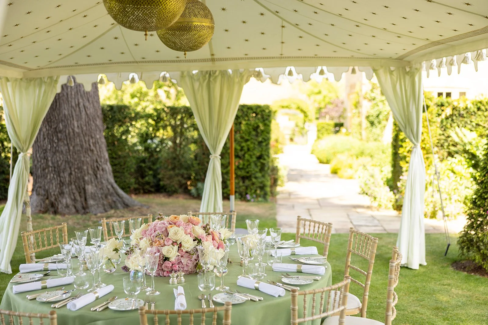 Outdoor event setup with a table under a canopy, decorated with a large floral centerpiece of pink and white flowers, surrounded by glassware, plates, and silverware. The canopy has cream curtains and hanging decorative balls, with trees, bushes, and a stone pathway visible in the background.