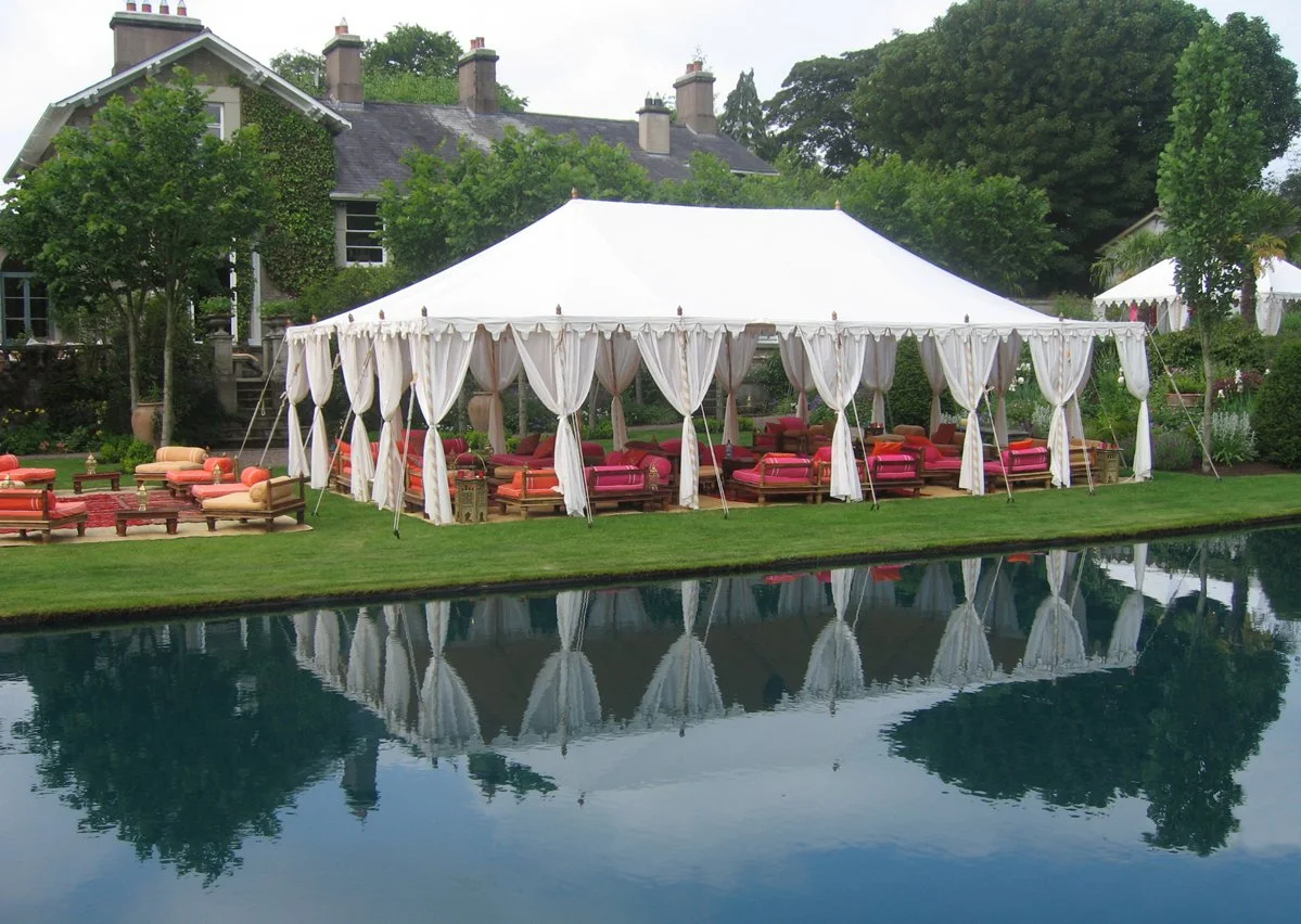 A large white event tent with white curtains, set up on a grassy lawn next to a body of water, with colorful chairs and sofas arranged inside and outside for seating. A house with chimneys and trees are in the background.