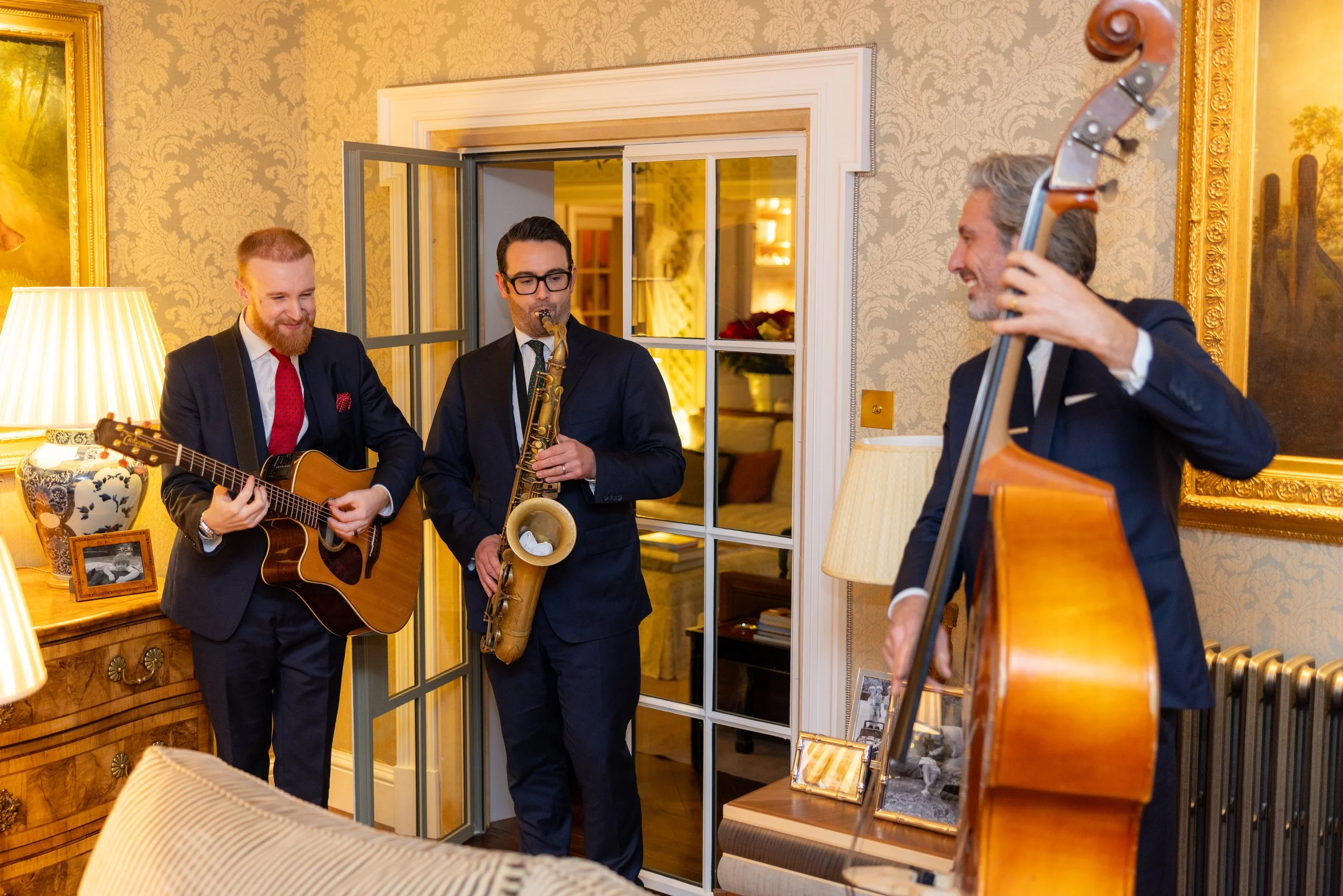 Three men in suits playing musical instruments in a warmly decorated room, with gold framed paintings and a glass display case, during what appears to be an indoor gathering.