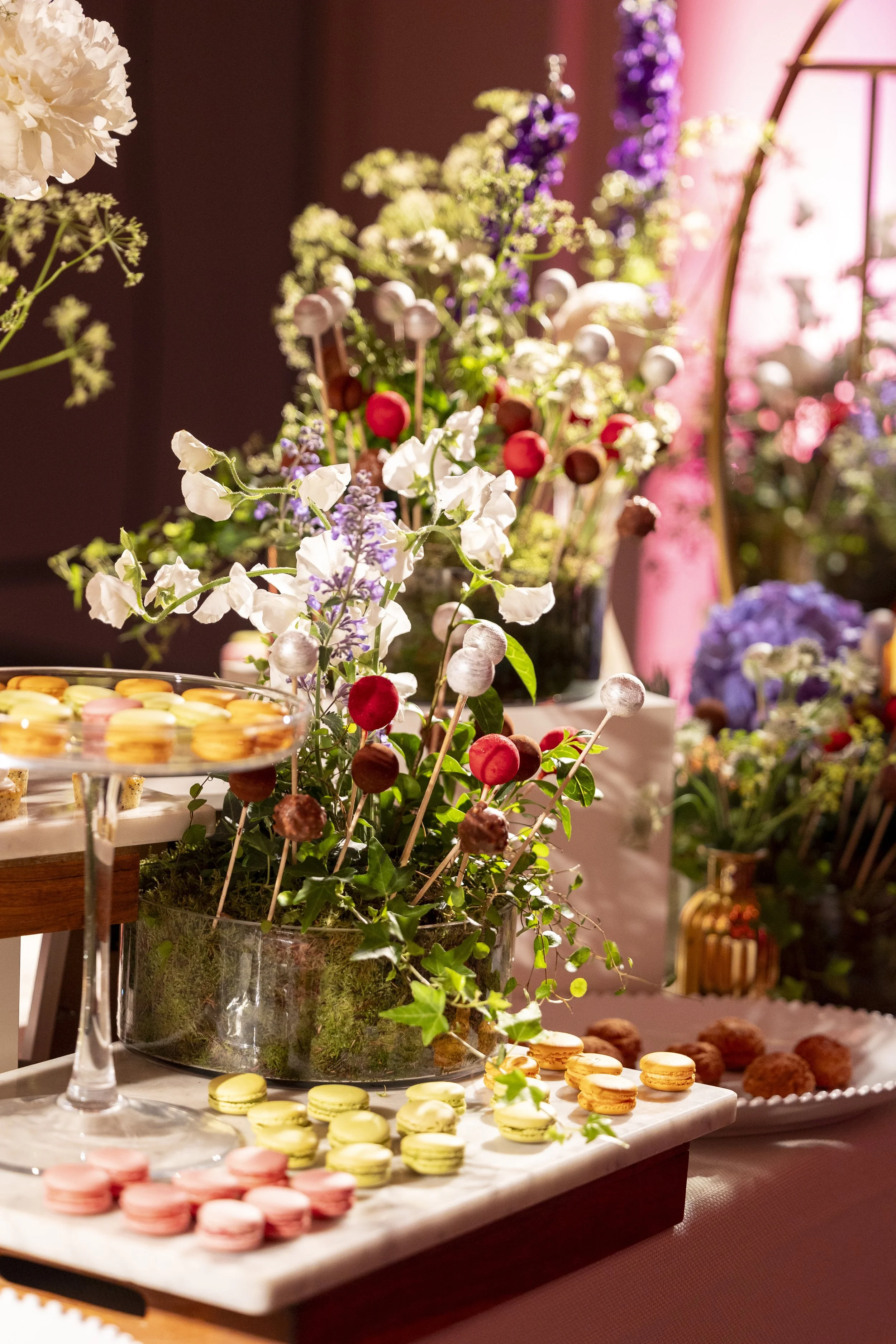 Close-up of a table with a variety of colorful macarons, chocolates, and floral arrangements in the background, set in a warmly lit indoor setting.