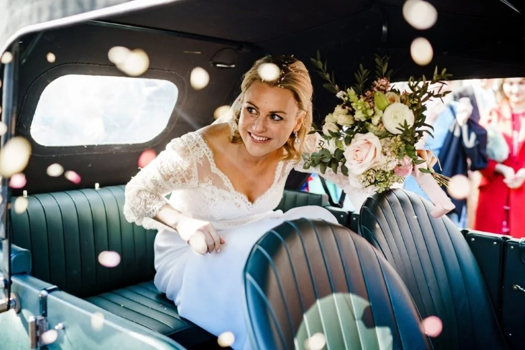 A bride in a white lace wedding dress sitting in the back of a vintage car, holding a large bouquet of flowers and smiling.