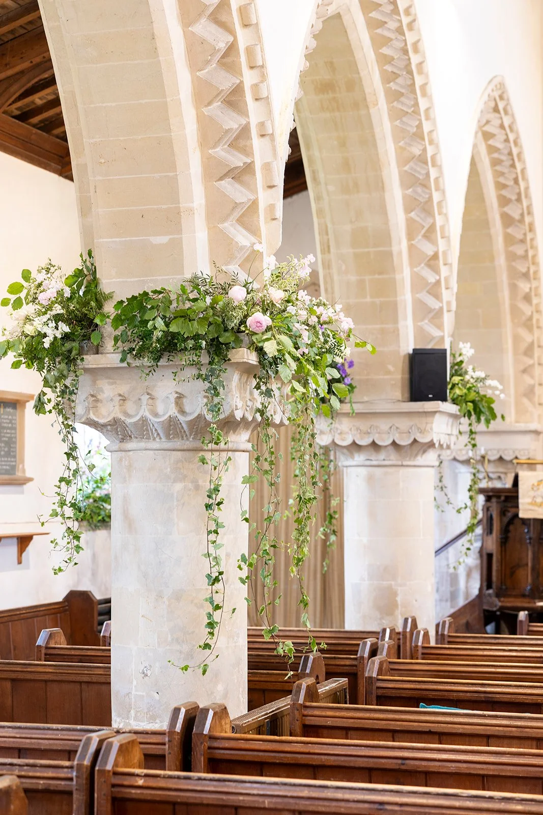 Interior of a church with tall stone pillars decorated with hanging green ivy and pink flowers, wooden pews, and a high arched ceiling.