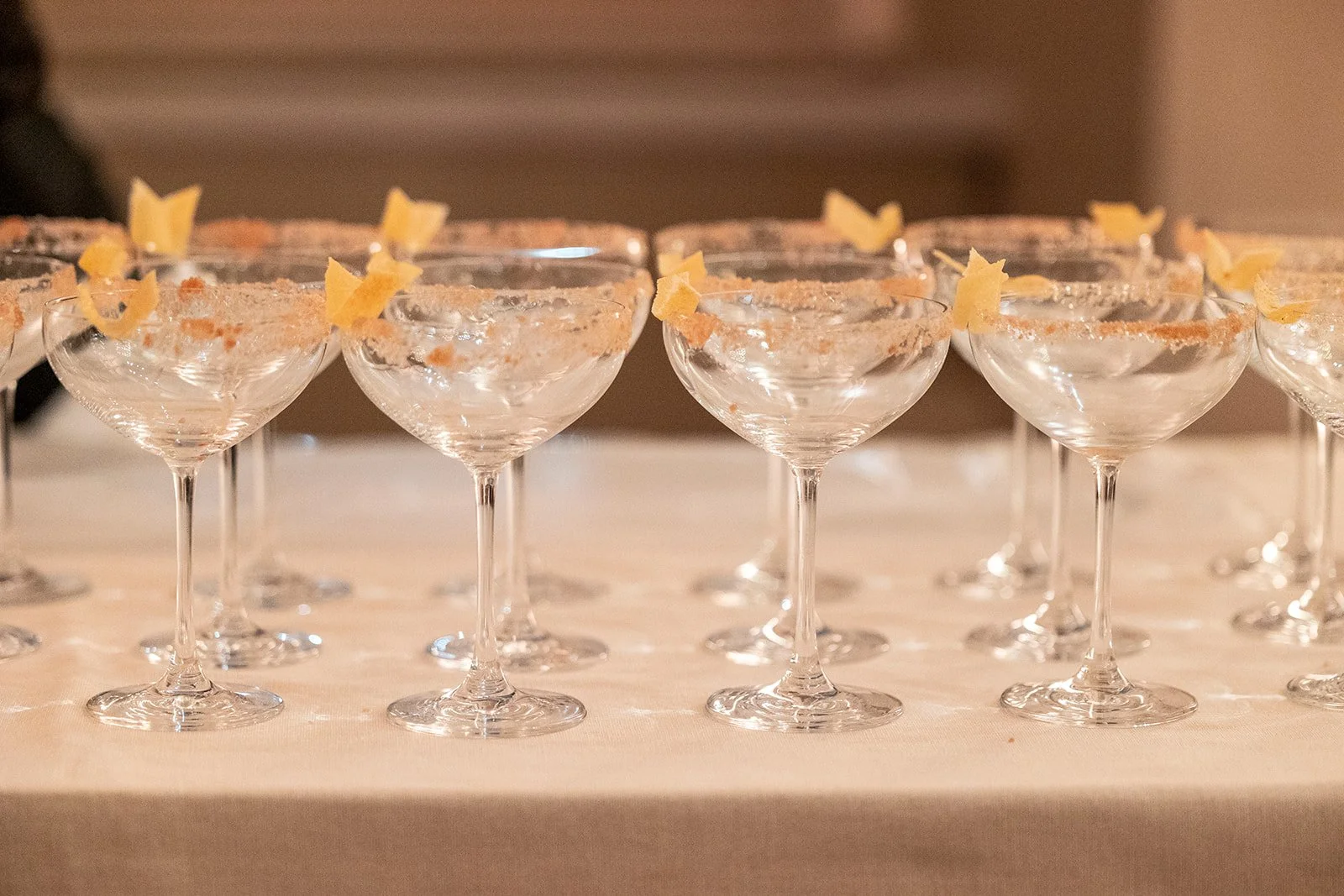 Multiple cocktail glasses with salted rims and garnished with lemon twists, arranged on a table for a party or celebration.