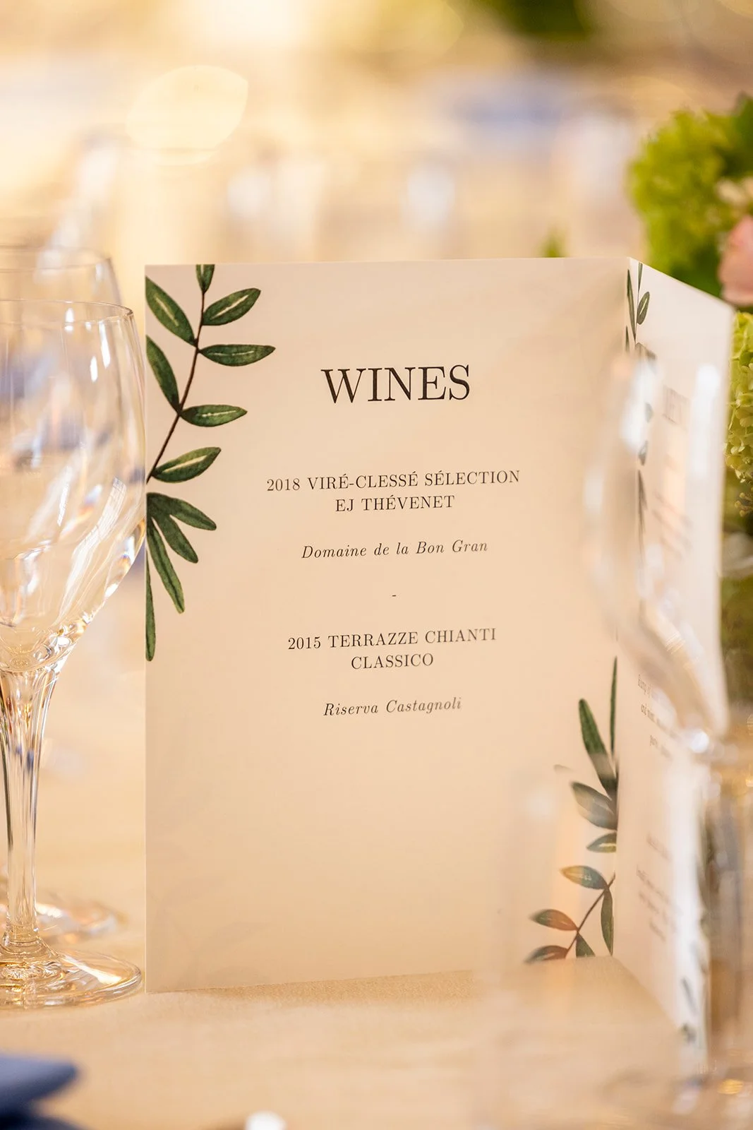 Menu sign featuring wine options with a decorative green leaf border, displayed on a table with wine glasses nearby at an elegant event.