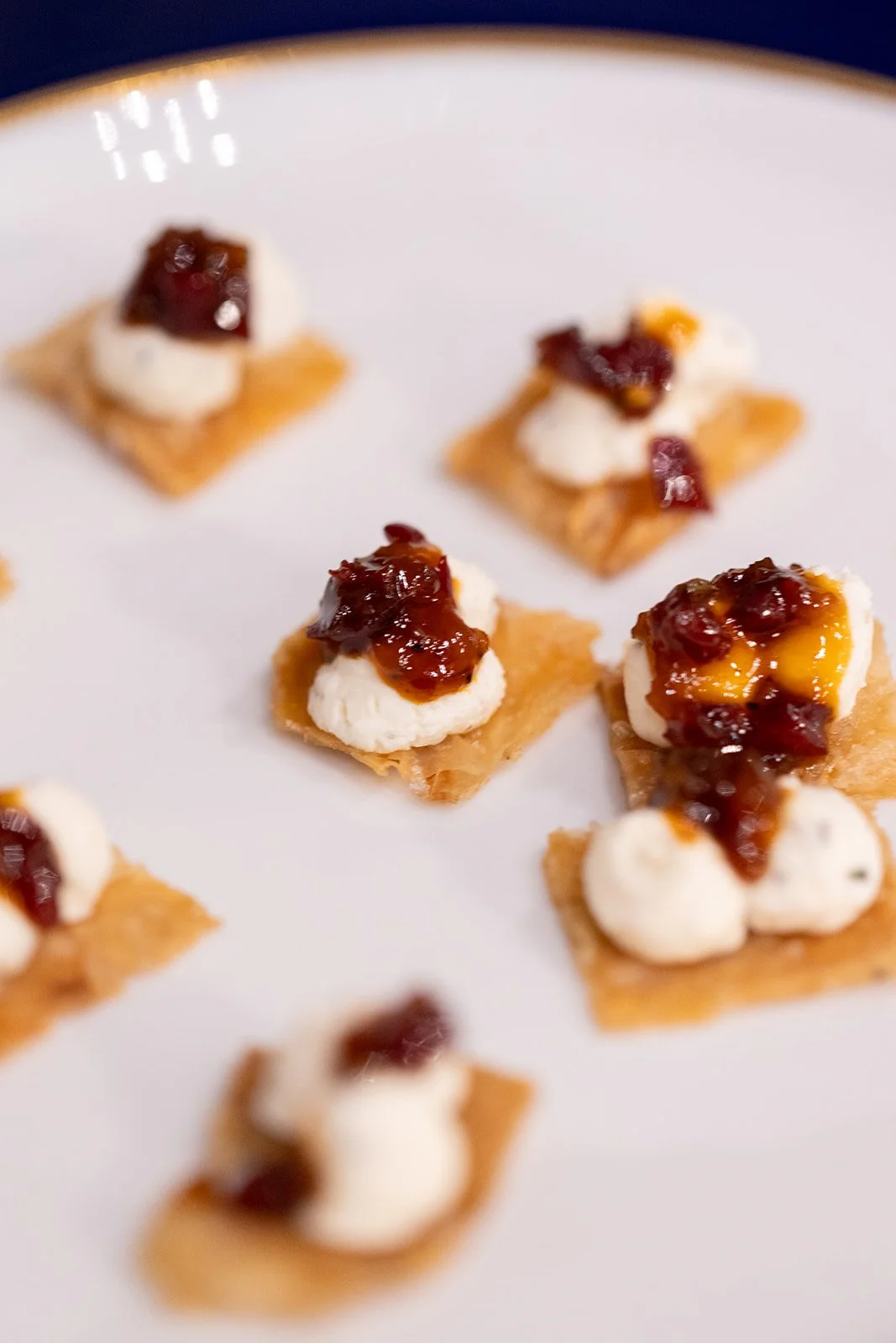 Mini appetizers with crackers topped with cream cheese, jam, and possibly fruit or nuts, arranged on a white plate.