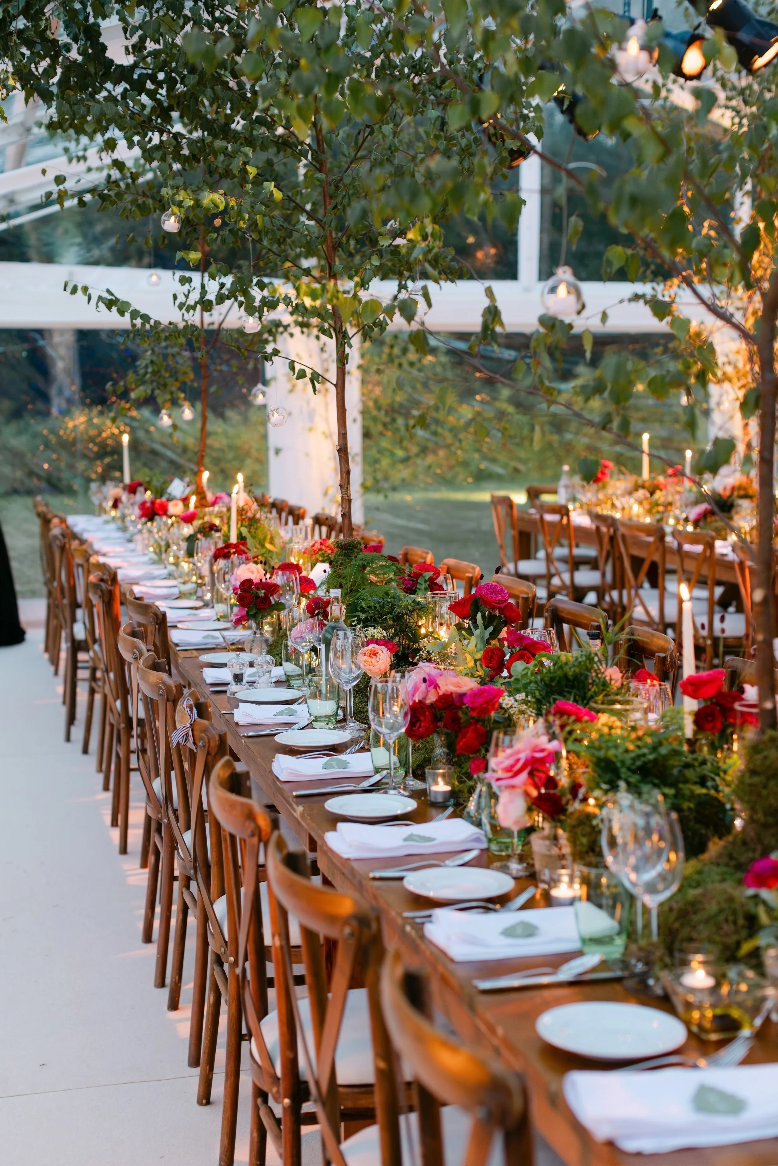 A long dining table decorated with flowers, candles, and glassware, set outdoors under string lights and surrounded by trees.