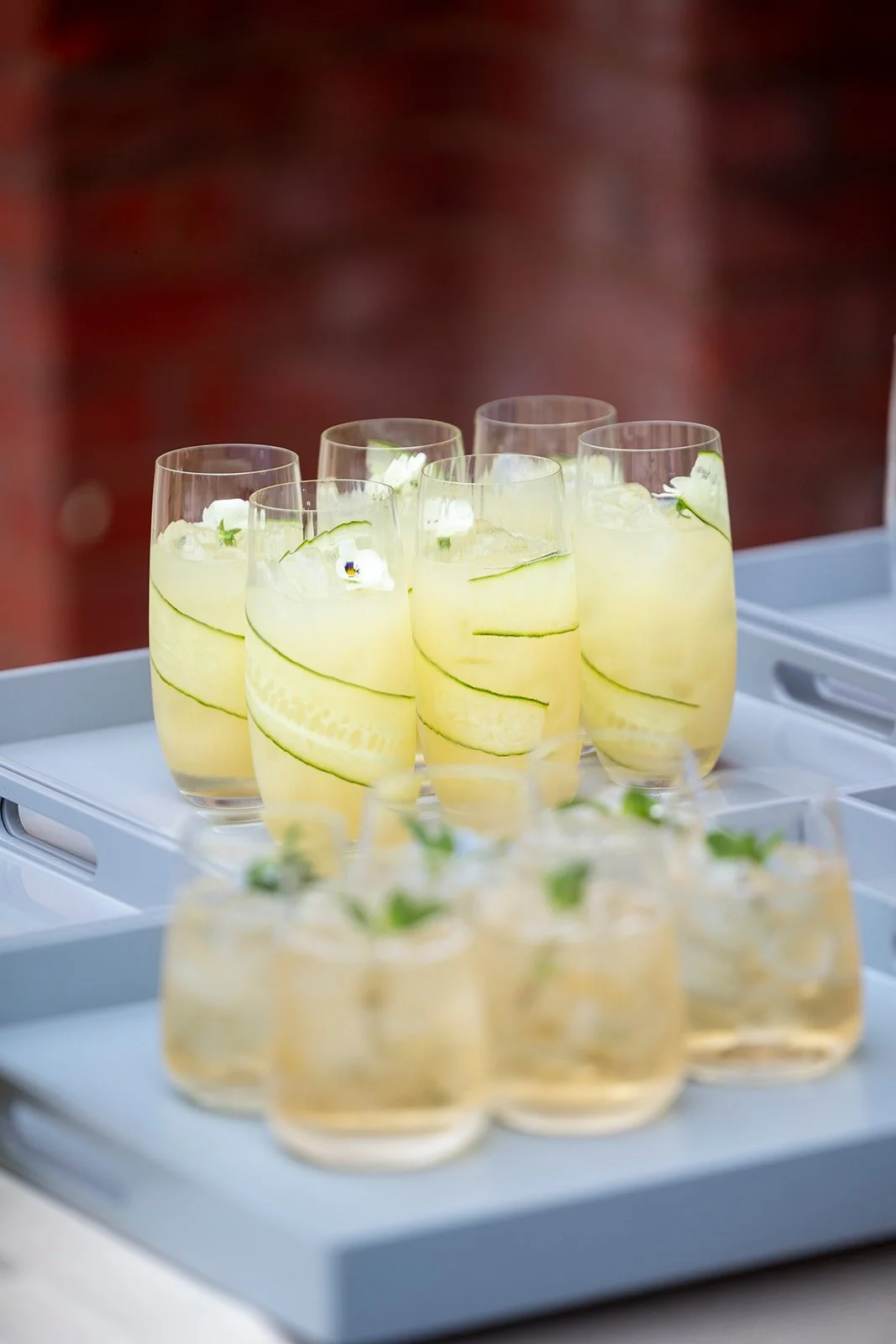 Glasses with a yellow drink, garnished with cucumber slices and small white flowers, on a serving tray.