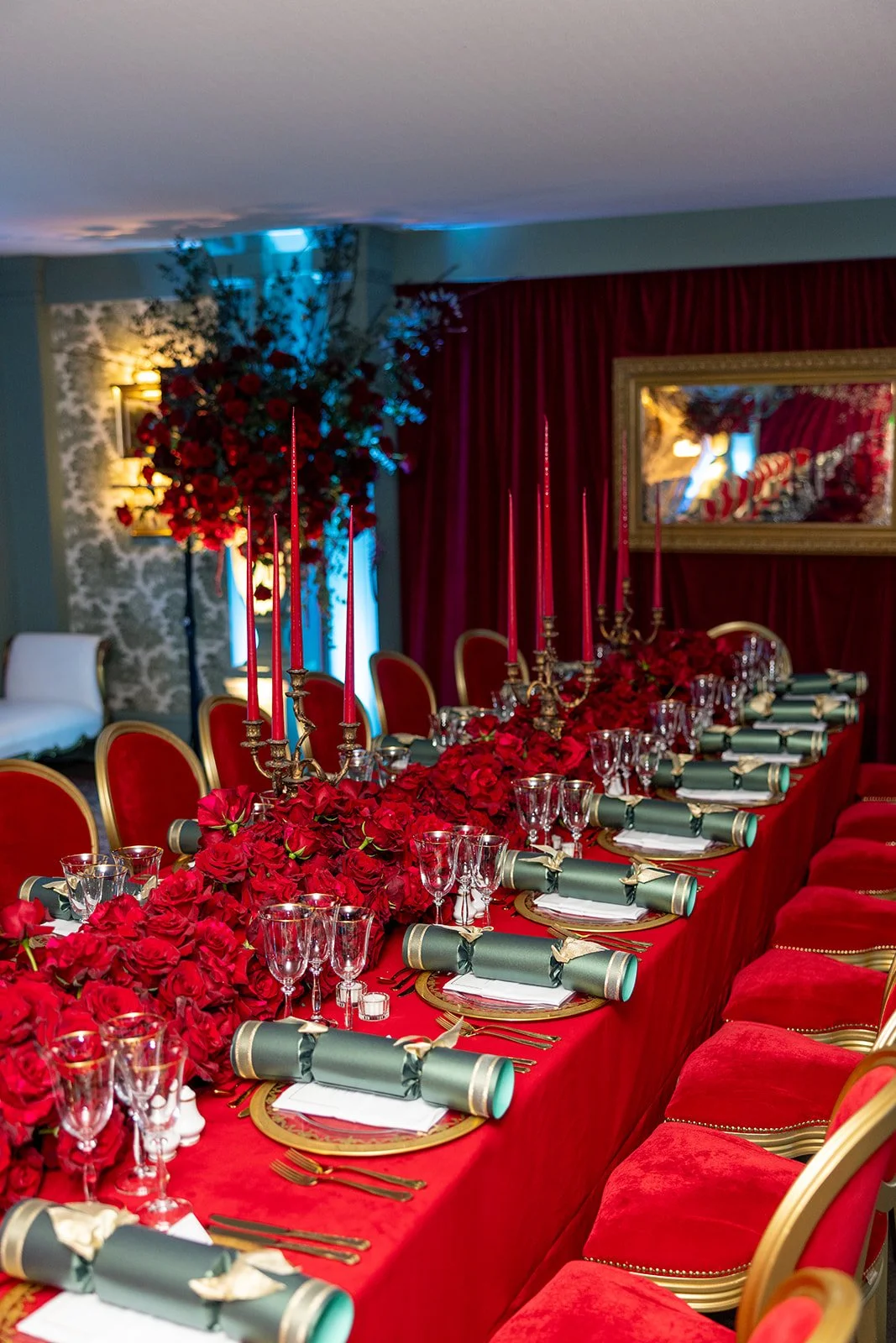 A luxurious dining table decorated with red roses, green napkins, gold cutlery, and tall red candles, set for a formal event in a decorated room with red curtains, floral wallpaper, and floral arrangements.