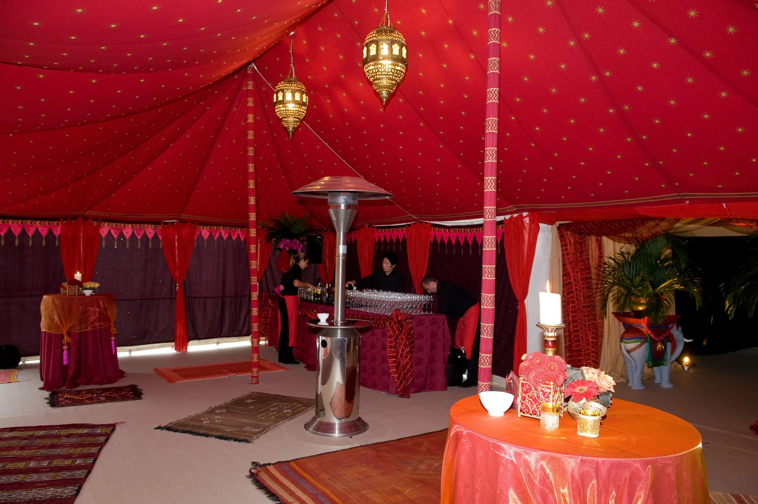 An event setup with a red tent or canopy decorated with gold stars, ornate hanging lanterns, and red drapes. Inside, there are tables with flowers, candles, and decorative items, with people preparing or organizing the space.