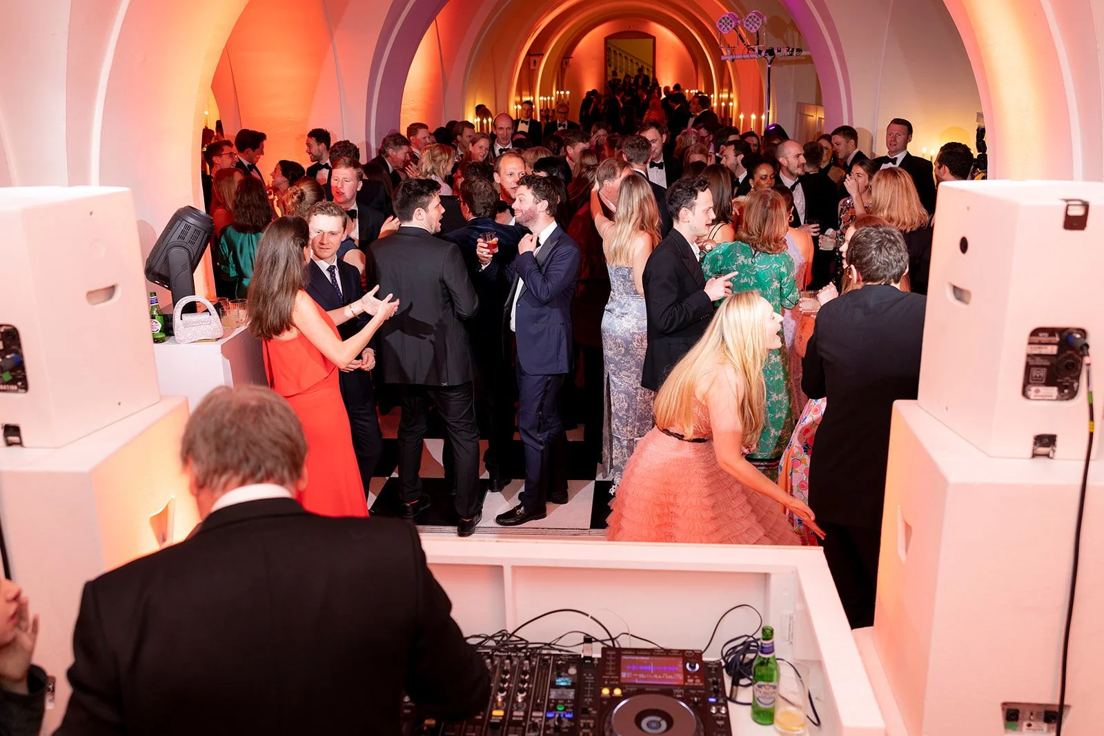 People dancing and socializing at a lively indoor party with colorful lighting, a DJ booth, and a crowd dressed in formal and semi-formal attire.
