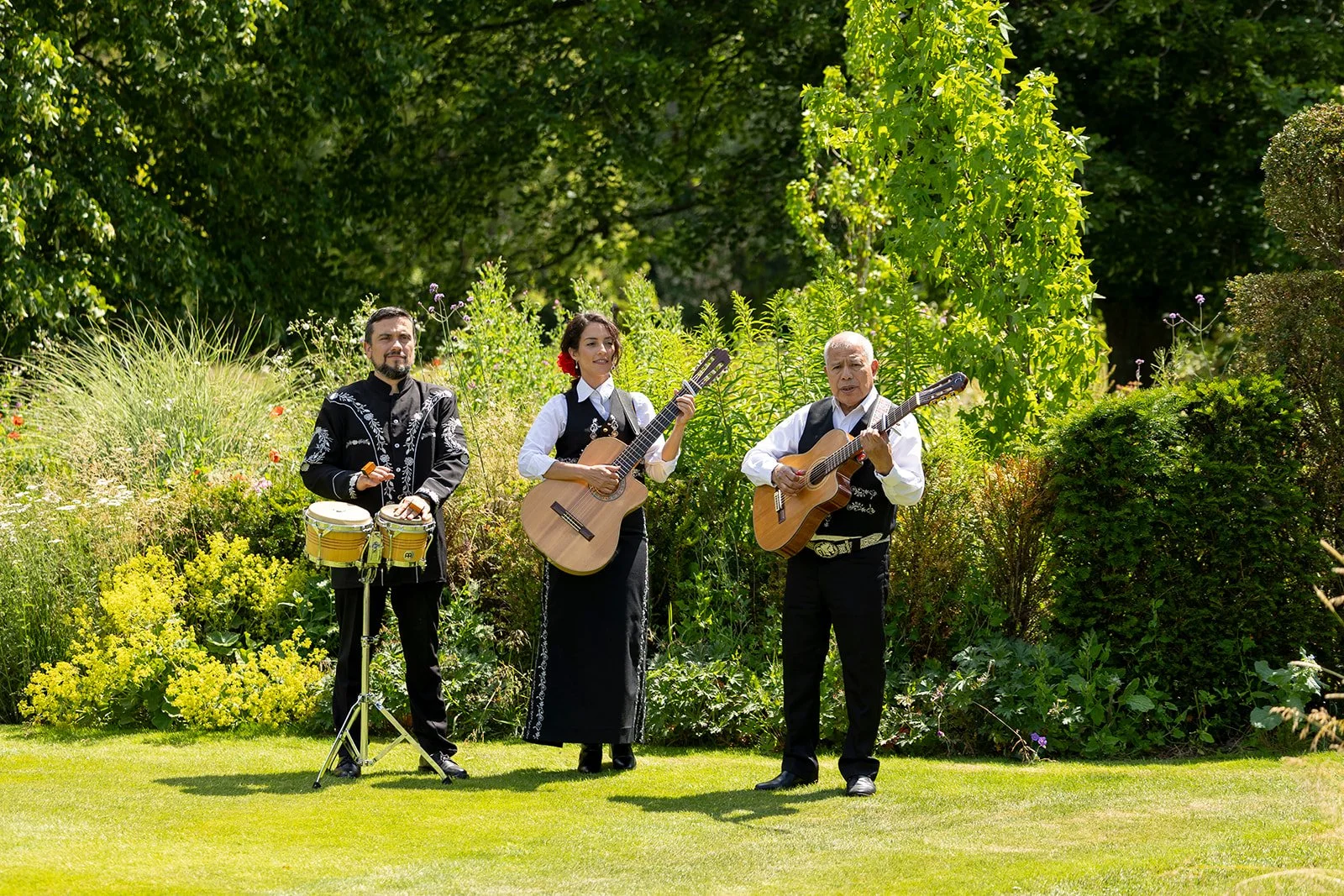 Three musicians playing instruments outdoors in a lush garden with green trees and shrubs in the background. The group includes two men and one woman, with the woman and one man playing guitars and the other man playing percussion.