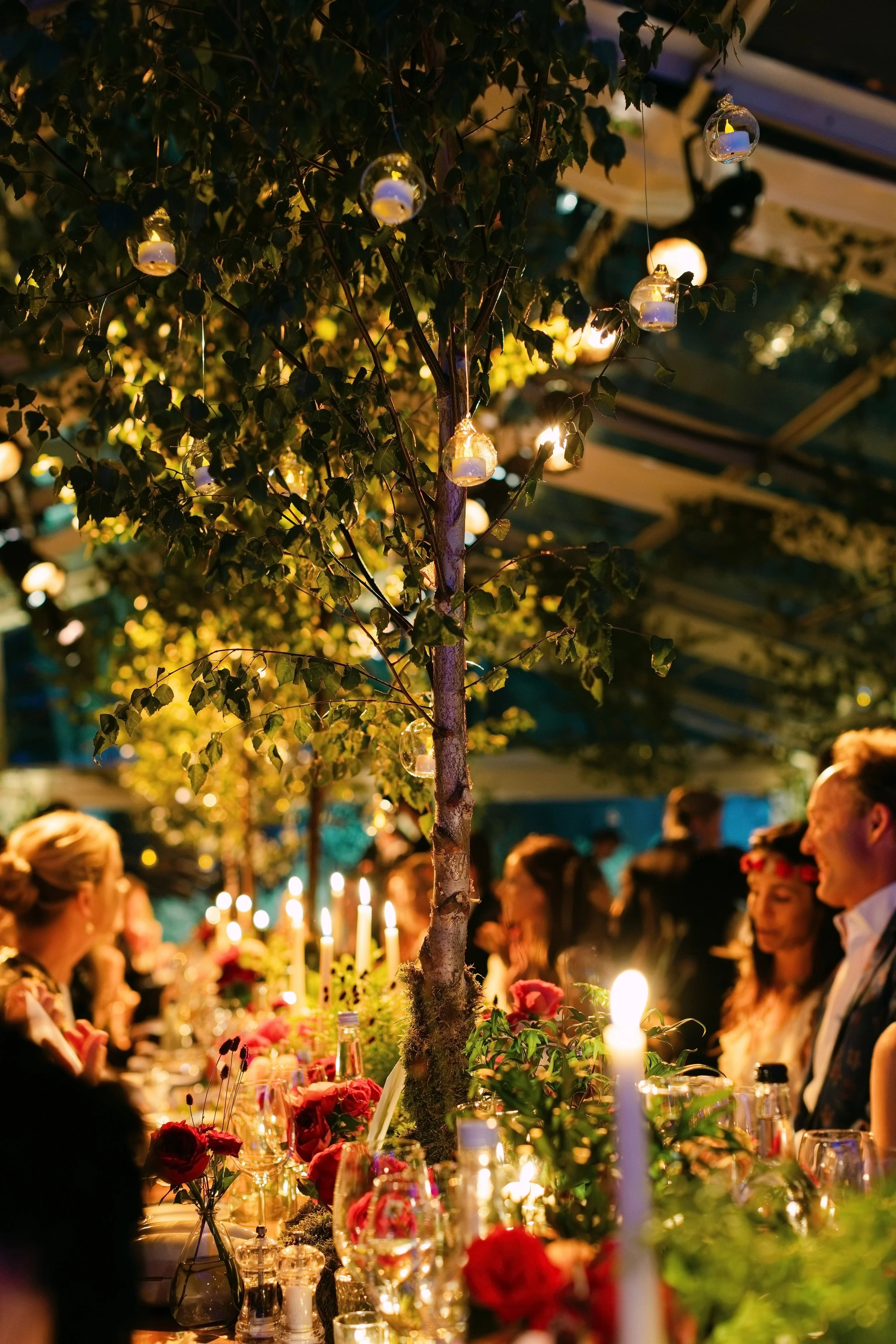 A nighttime indoor celebration with a long table decorated with candles, flowers, and greenery, surrounded by people socializing and enjoying the event.