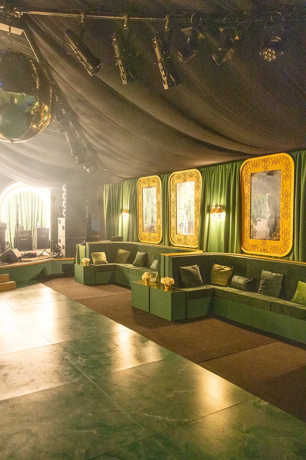 Empty lounge with green sofas, gold framed mirrors, green curtains, and stage with musical equipment at the back.