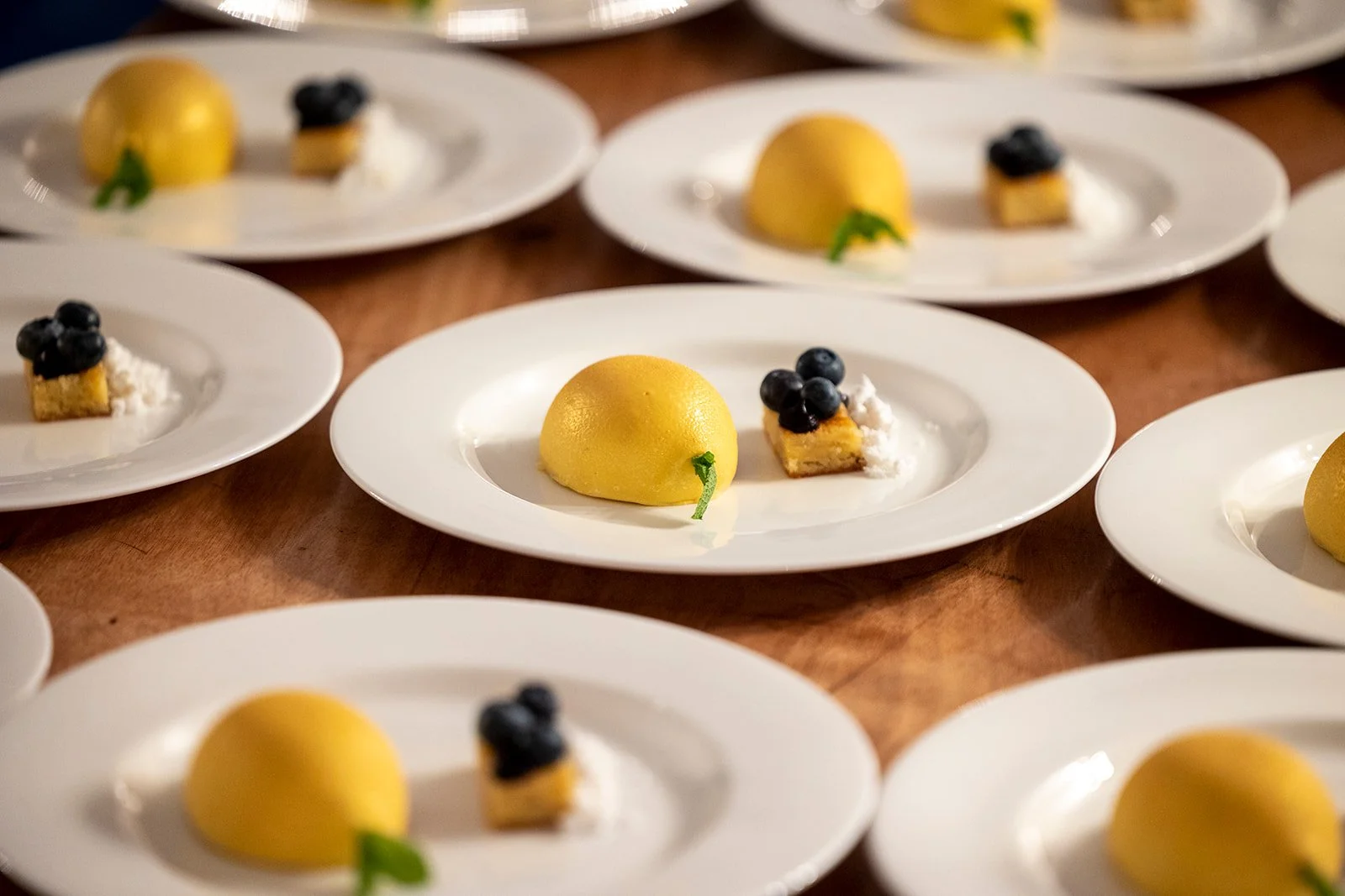 Multiple plates with yellow dome-shaped desserts, garnished with small green leaves, paired with small rectangular pastry pieces topped with blueberries and white cream, arranged on a wooden surface.