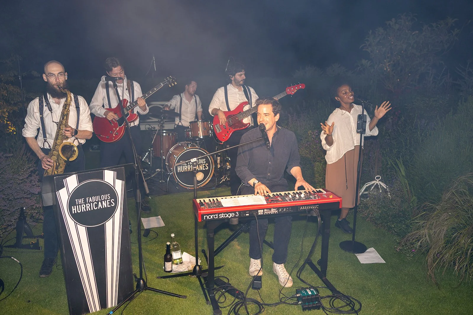 A band performing outdoors at night, featuring a keyboardist singing into a microphone, a singer with a microphone, a guitarist, a bassist, a drummer, and a saxophonist. There is a sign that reads 'The Fabulous Hurricanes'.