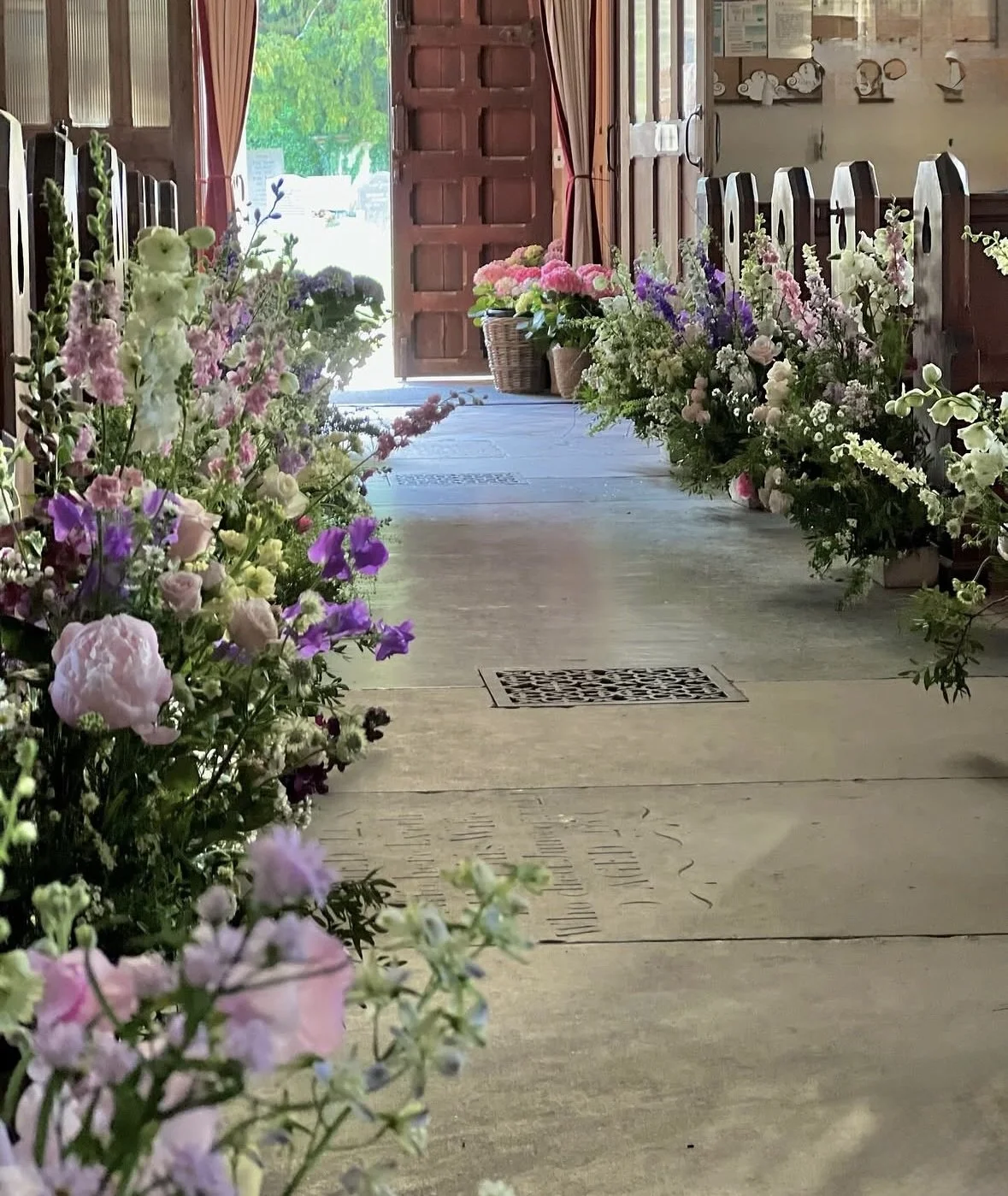 Flower arrangements line the sides of an indoor hallway with open wooden door at the end, leading outside to green trees and bright light.