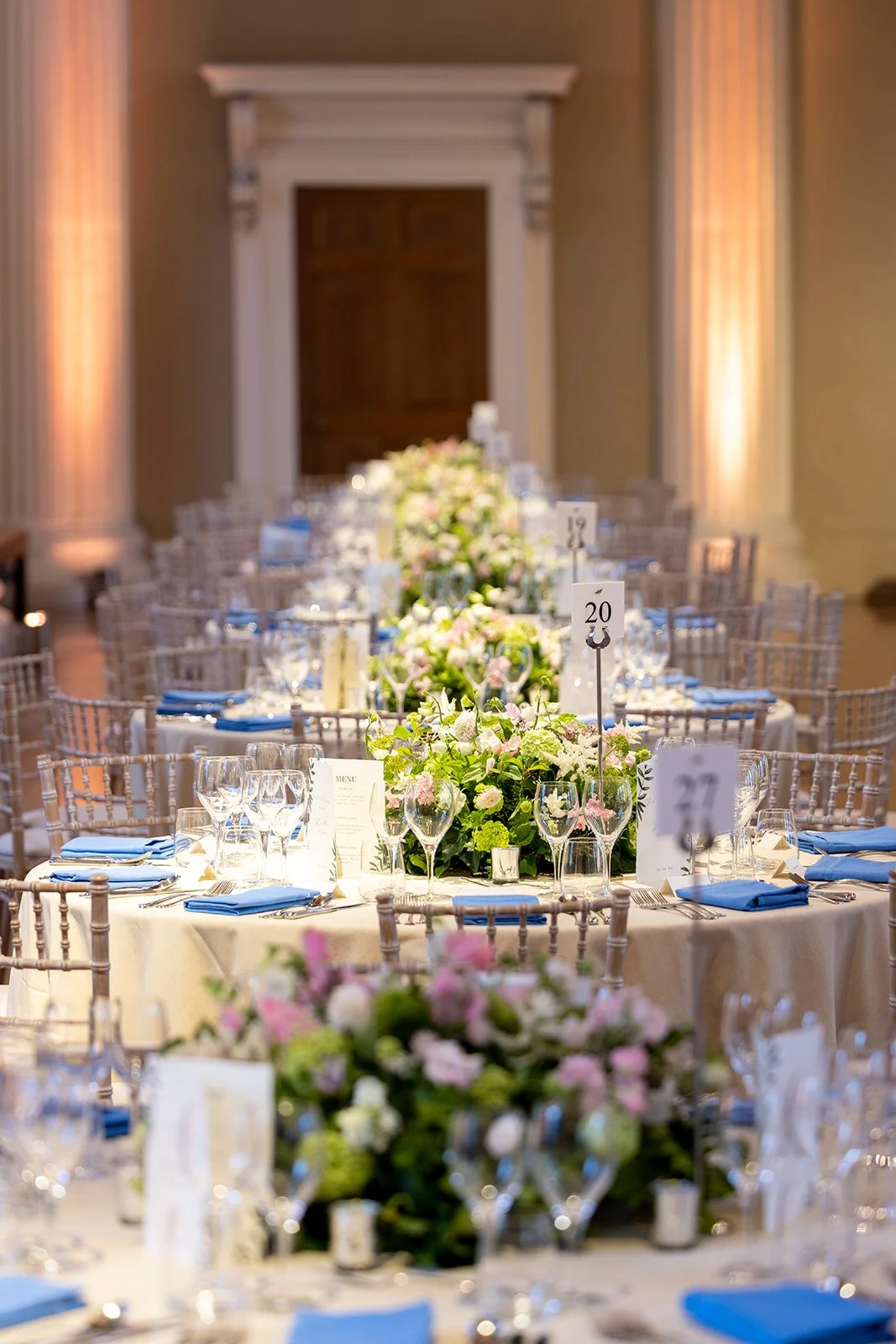 Elegant banquet table at a wedding or formal event, decorated with floral centerpieces, glassware, blue napkins, and table numbers, in a grand room with high ceilings and wooden door.