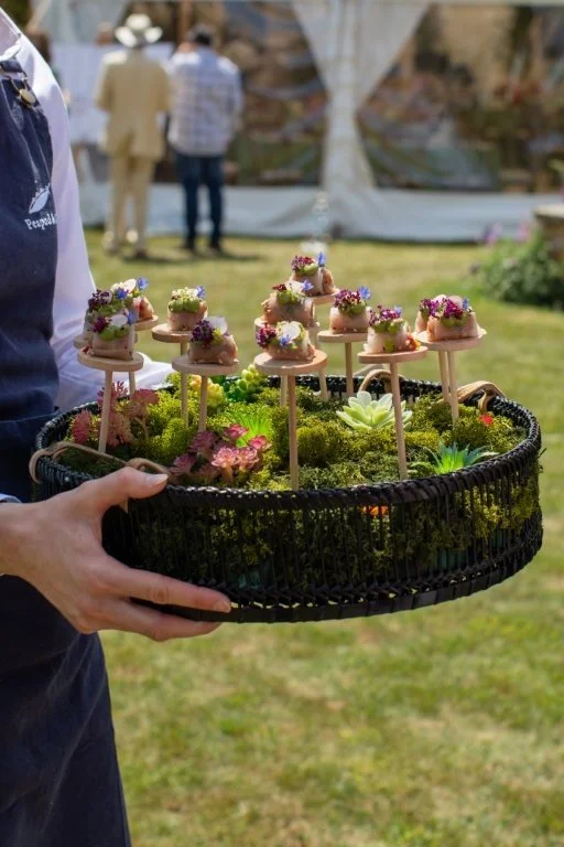 Person holding a black basket with small appetizers decorated with flowers on sticks, placed on moss with small plants, outdoors at a casual event.