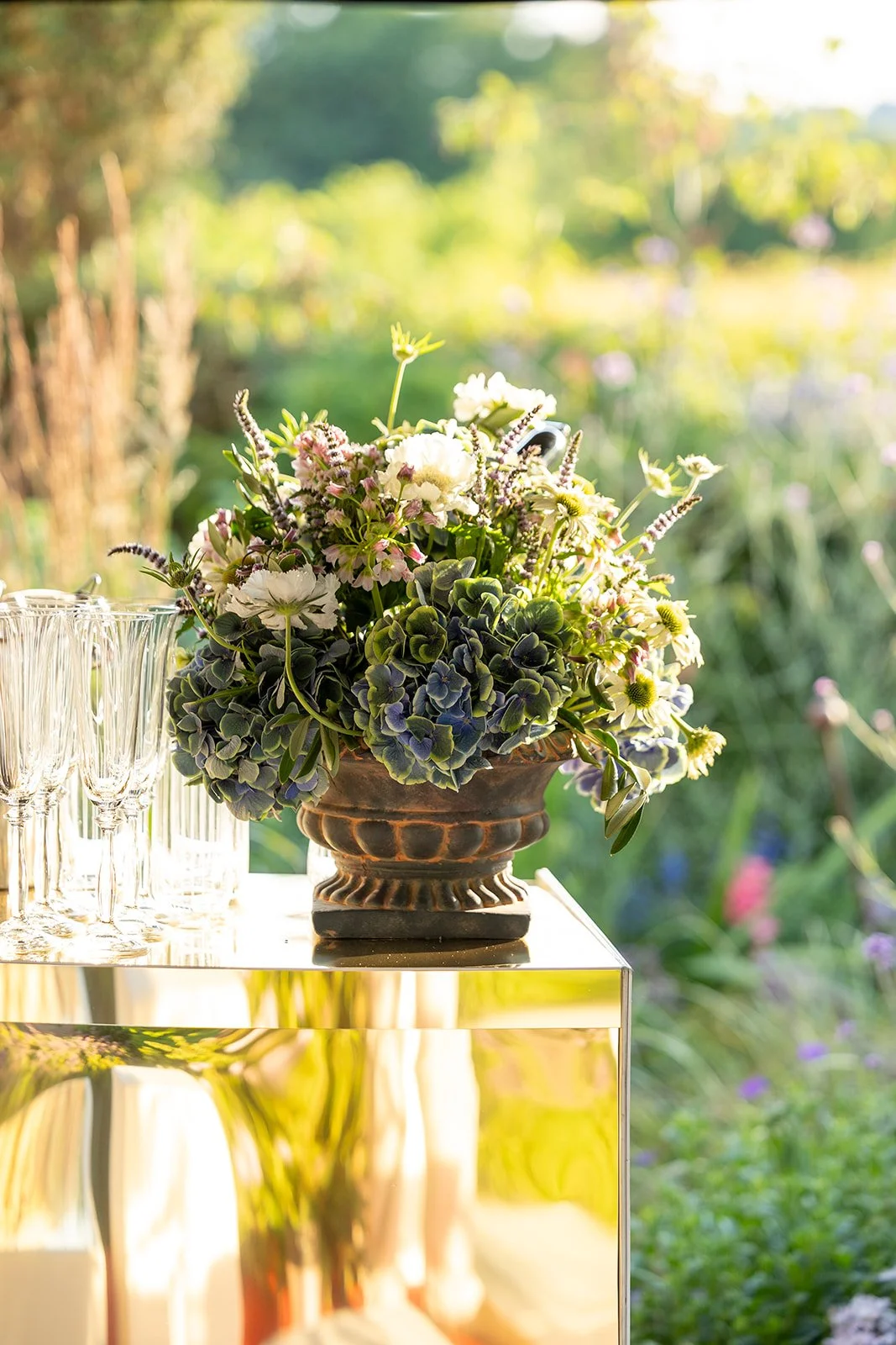 A bouquet of white and purple flowers in a bronze vase on a reflective surface outdoors, with a blurred garden background.