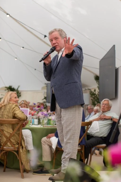 A man in a blue blazer and gray pants speaking into a microphone at a social event, with attendees seated at tables behind him under a white tent.