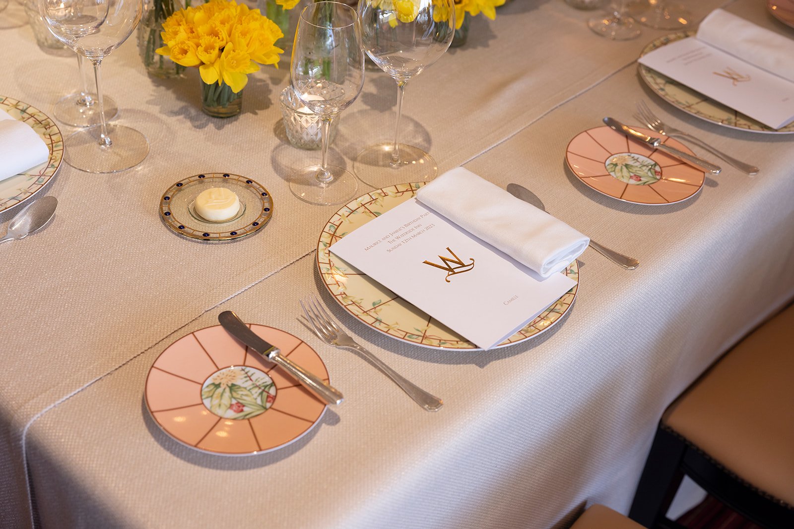 A table set for a formal event with floral plates, silverware, wine glasses, and a menu card. There are yellow daffodils in the background and a lit candle in the center.