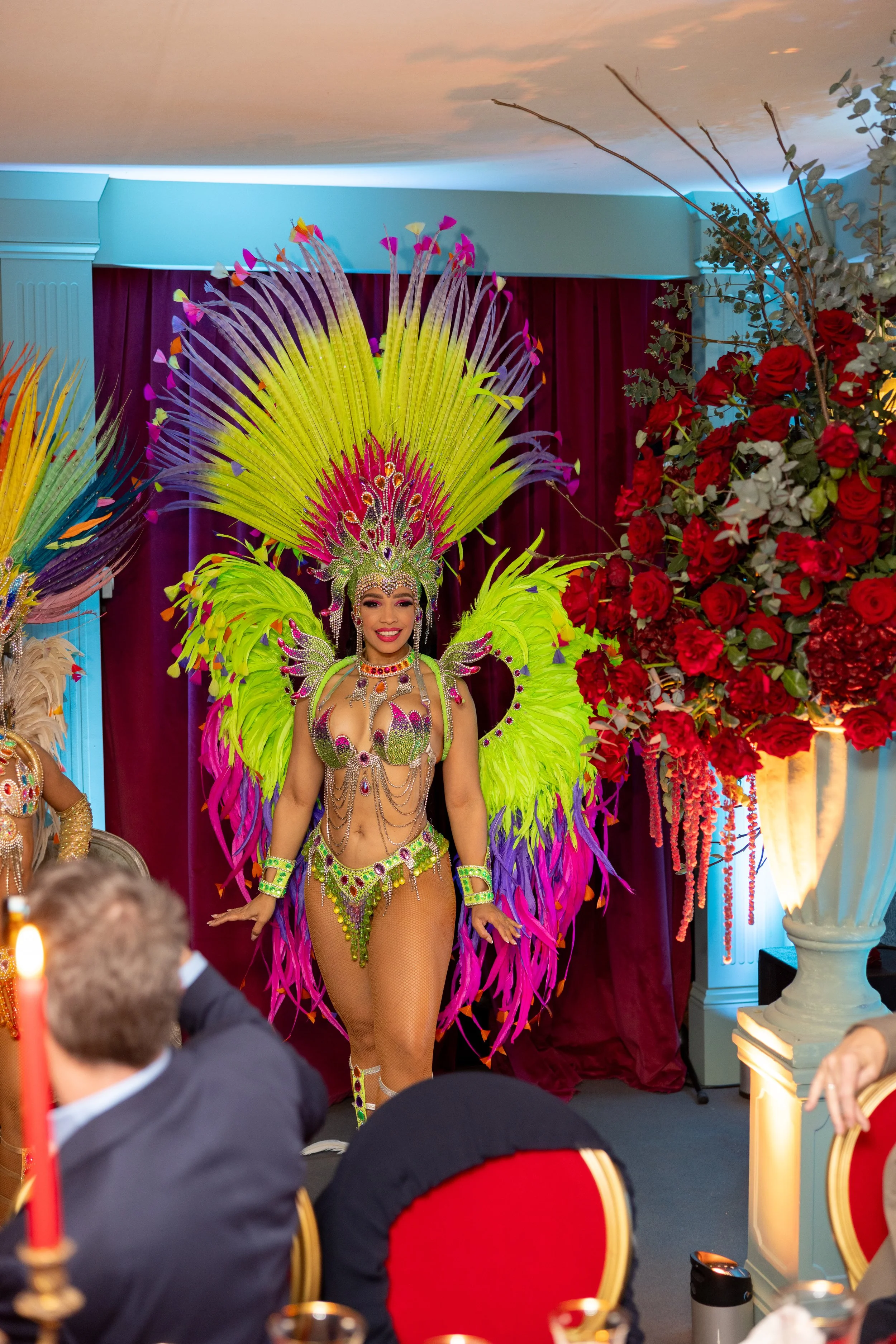 A woman in colorful carnival costume with large feather headdress and jewelry, smiling as she walks down a runway at an indoor event with a dark red curtain and floral arrangement in the background.