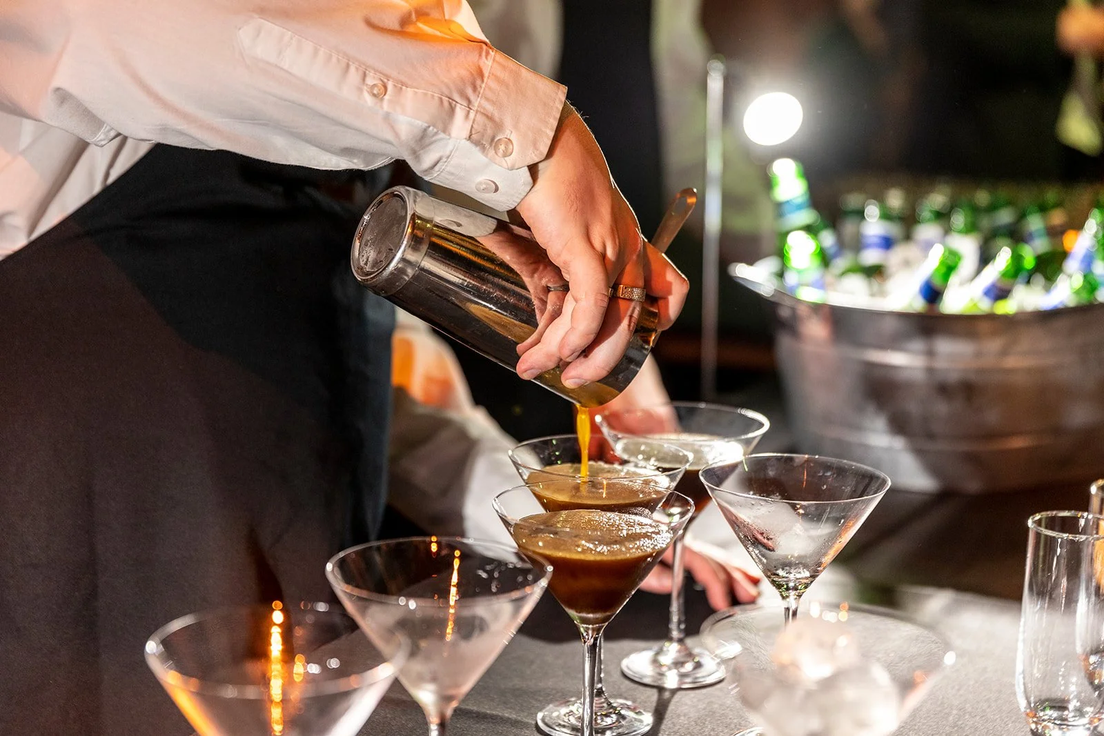 A person pours a dark cocktail into a martini glass at a bar or party setting, with several empty martini glasses on the table and a large container of bottles in the background.