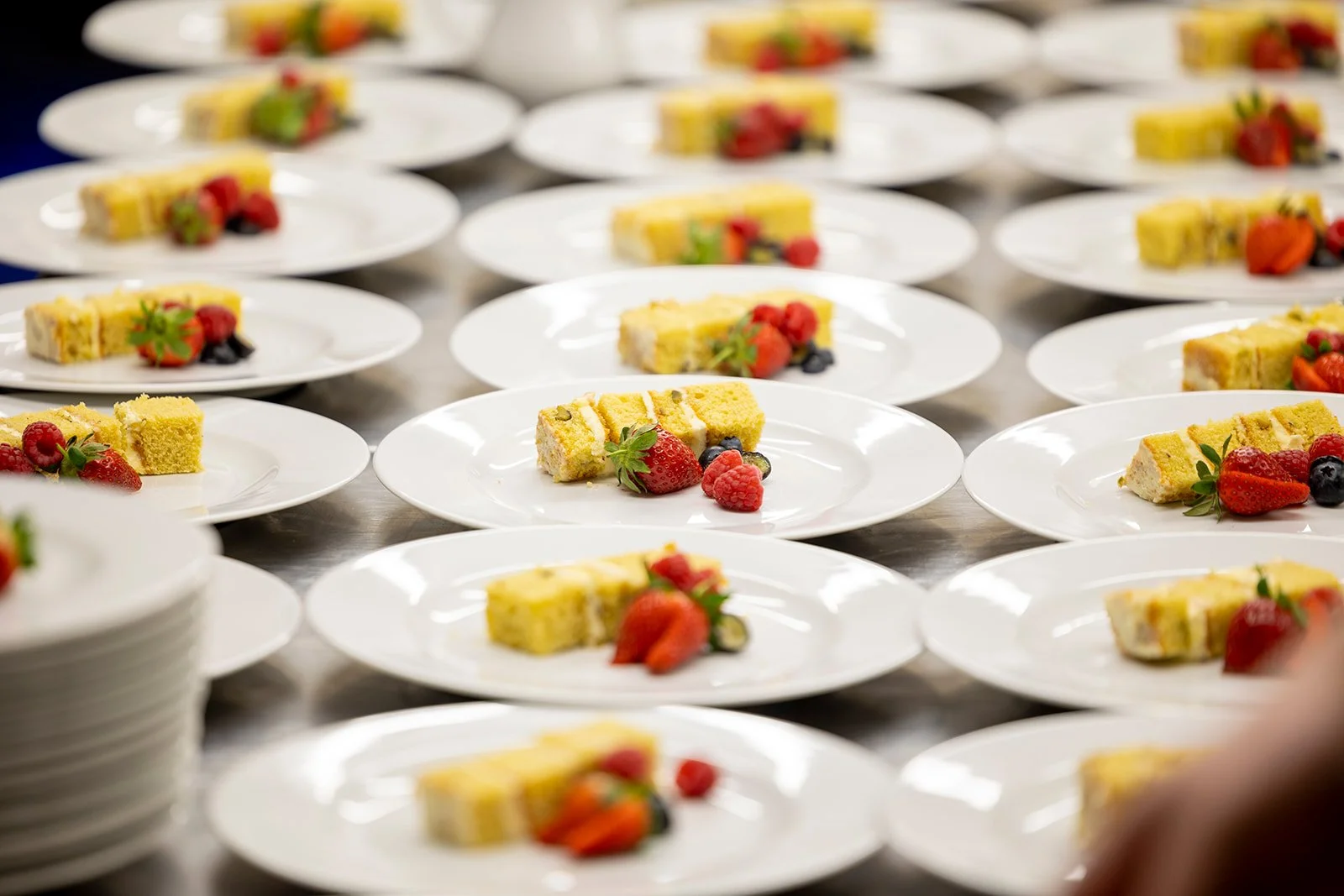 Multiple white plates with slices of layered cake topped with berries and garnished with mint, arranged on a busy service table.