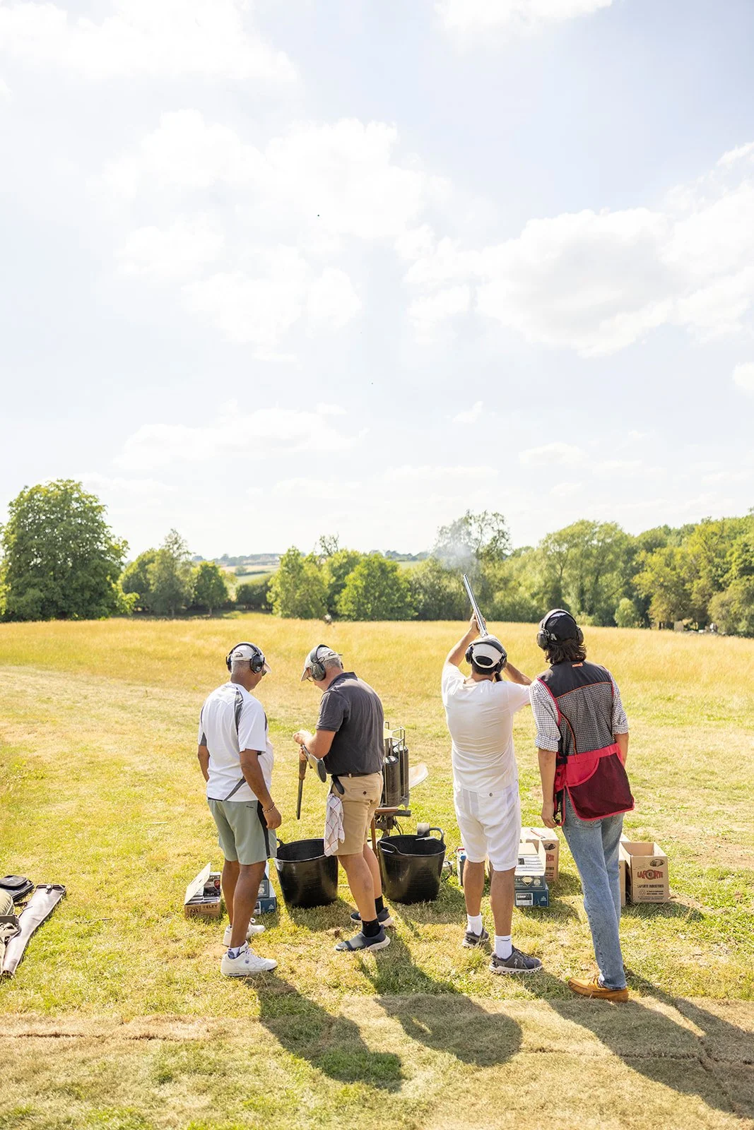 Four men at a shooting range with guns, targets, and protective gear on a sunny day.