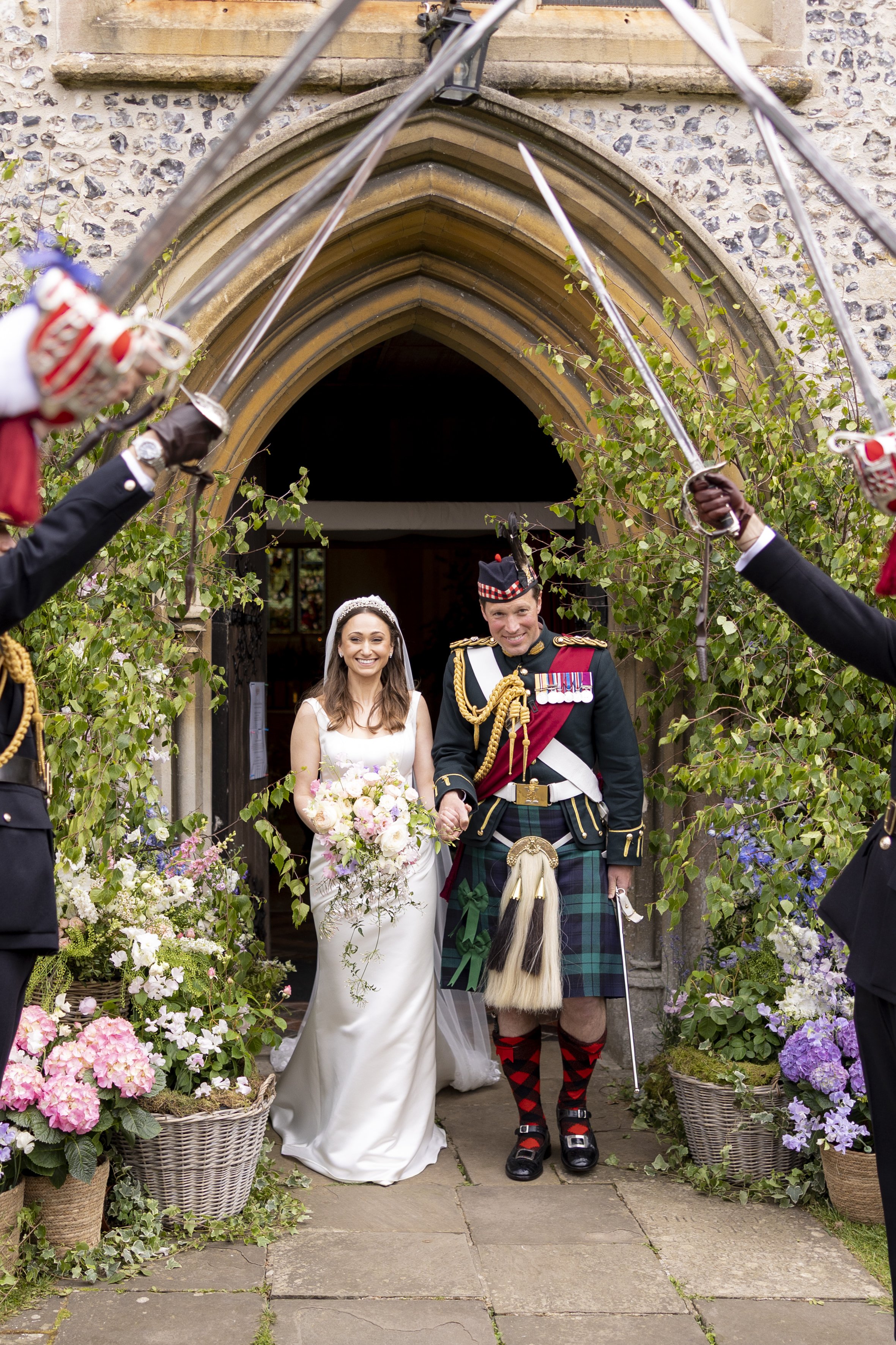 A bride and groom, dressed in wedding attire, walking out of a church or chapel while flower soldiers hold swords above them. The bride is wearing a white wedding dress and holding a bouquet, and the groom is wearing a traditional Scottish kilt with 