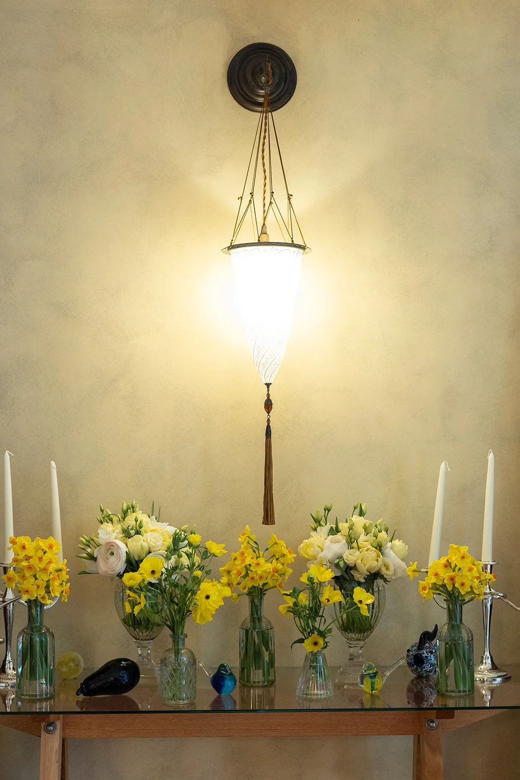 Table with flower arrangements in glass vases, white taper candles, and decorative glass animals, illuminated by a hanging ceiling light fixture.