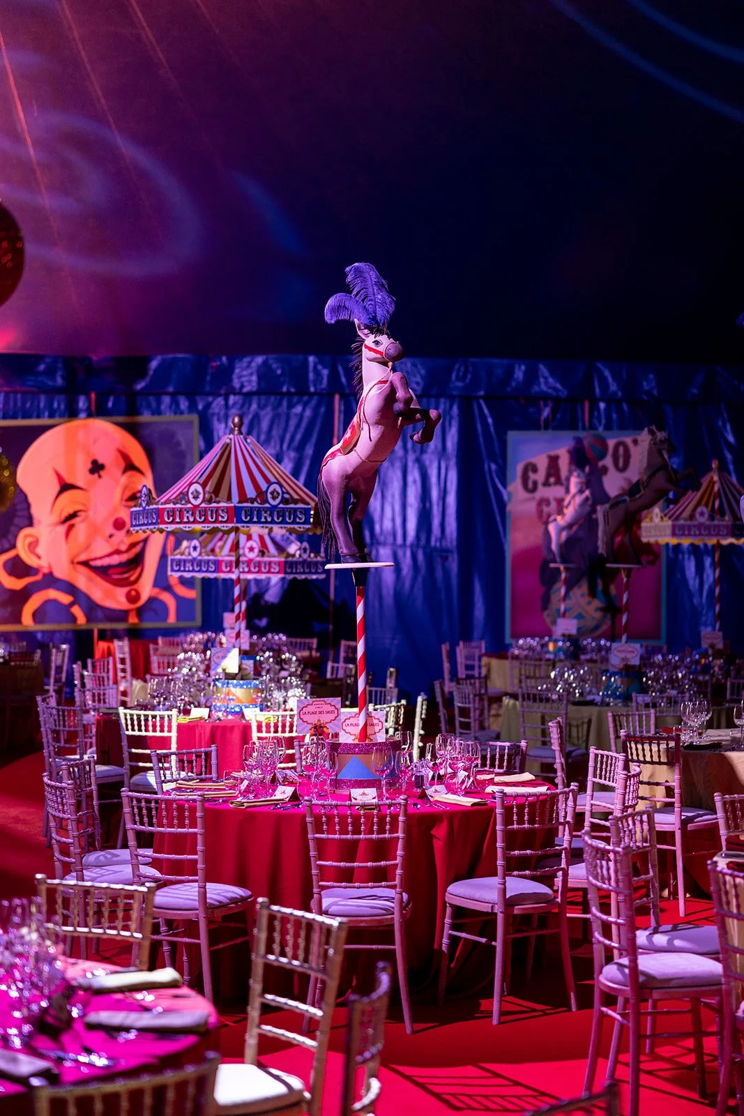 Inside a circus-themed event tent with round tables covered in pink tablecloths, set with glassware and silverware, surrounded by white chairs. A central decorative element features a tall striped pole with a mounted cartoonish horse with a purple fe