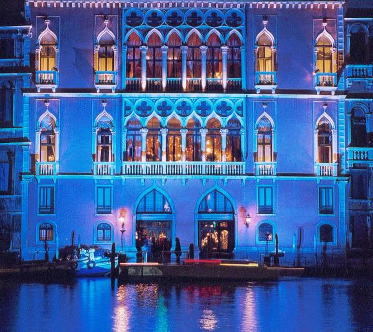 A brightly lit multi-story building illuminated in blue with Gothic architectural design, including pointed arches and ornate tracery, reflected on a water surface in the foreground.