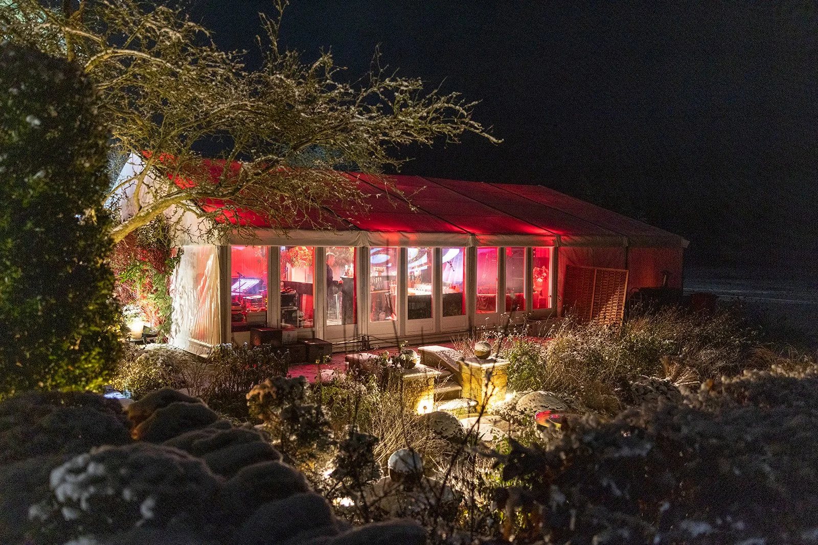 A nighttime scene of a red tent with glass walls, illuminated from within, set among plants and rocks, with snow on the ground and trees nearby.