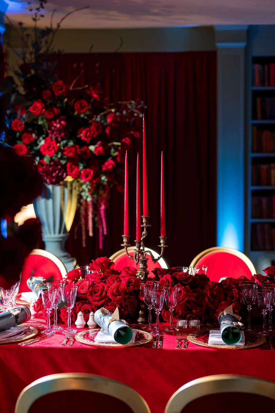 Elegant table setting with red roses and tall red candles, surrounded by gold-rimmed glassware and white napkins, set against a backdrop of red curtains and a large floral arrangement.