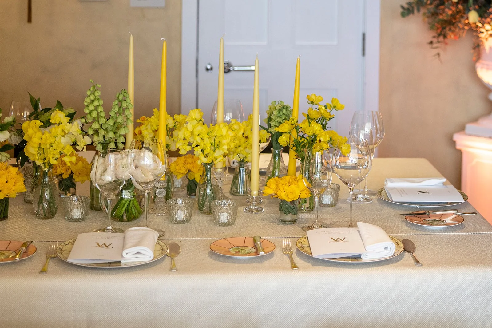 Elegant dinner table set with yellow flowers, tall yellow candles, glassware, plates, and silverware on a beige tablecloth.