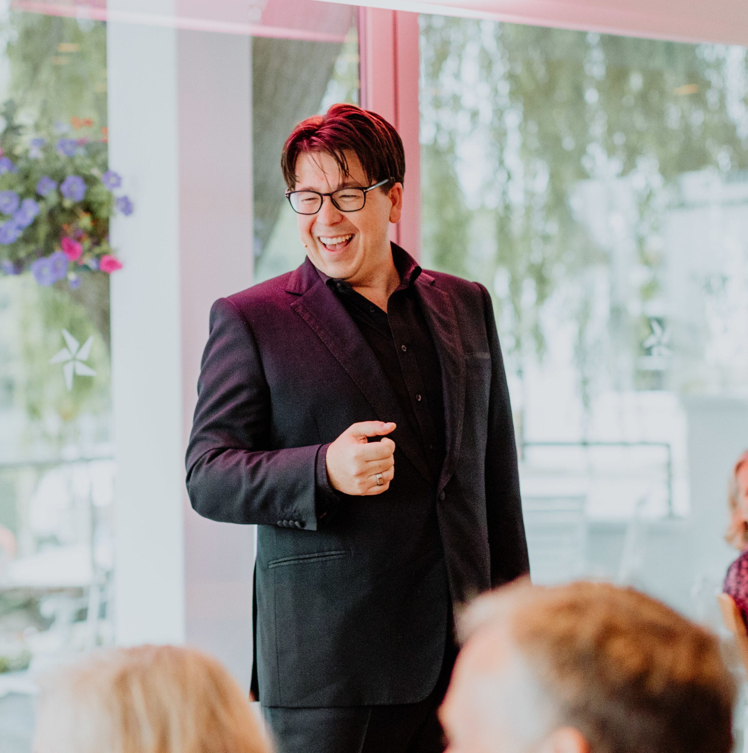 Smiling man with red hair, glasses, wearing a dark blazer, standing near a glass window with trees outside.