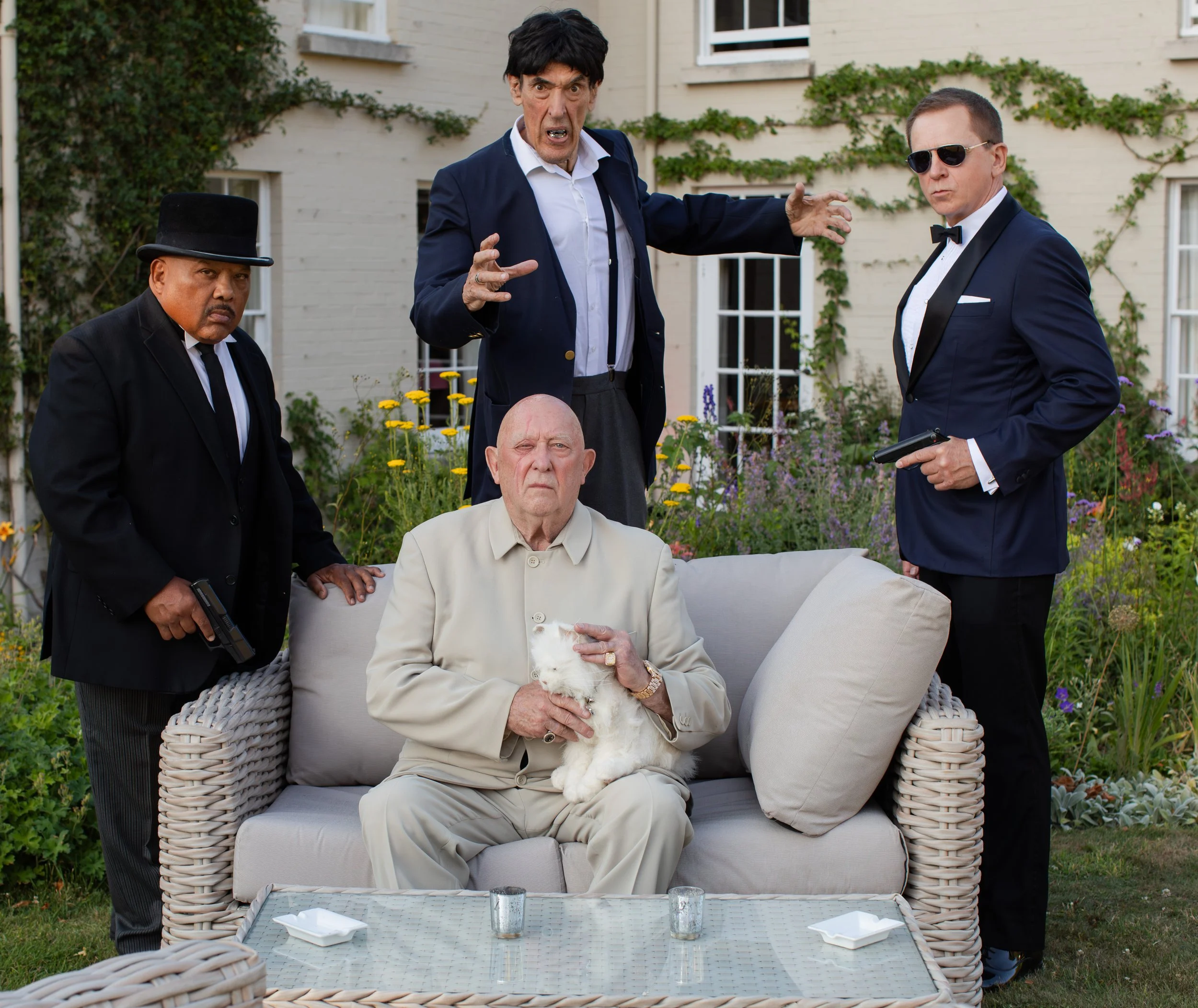 A group of men in formal attire, including tuxedos and sunglasses, gathered outdoors near a building with greenery. One man in a beige suit is sitting on a wicker sofa holding a white fluffy dog, while another man in a blue suit and black wig appears