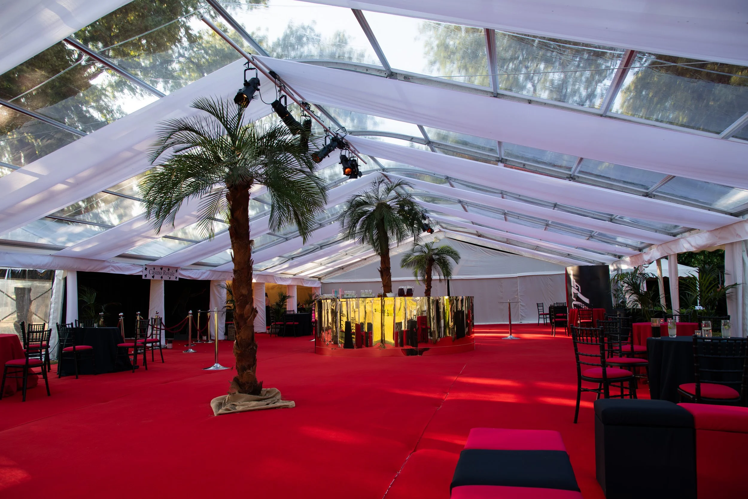 An event space with red carpet flooring, decorated with palm trees, black chairs with red cushions, and a gold mirror wall. The ceiling is made of glass panels with draped white fabric and stage lighting.