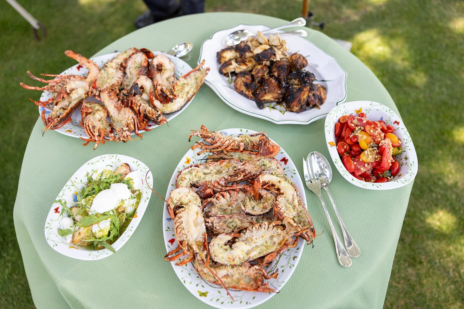 Seafood feast with lobster, grilled chicken, salad, and cherry tomato salad on a green tablecloth.
