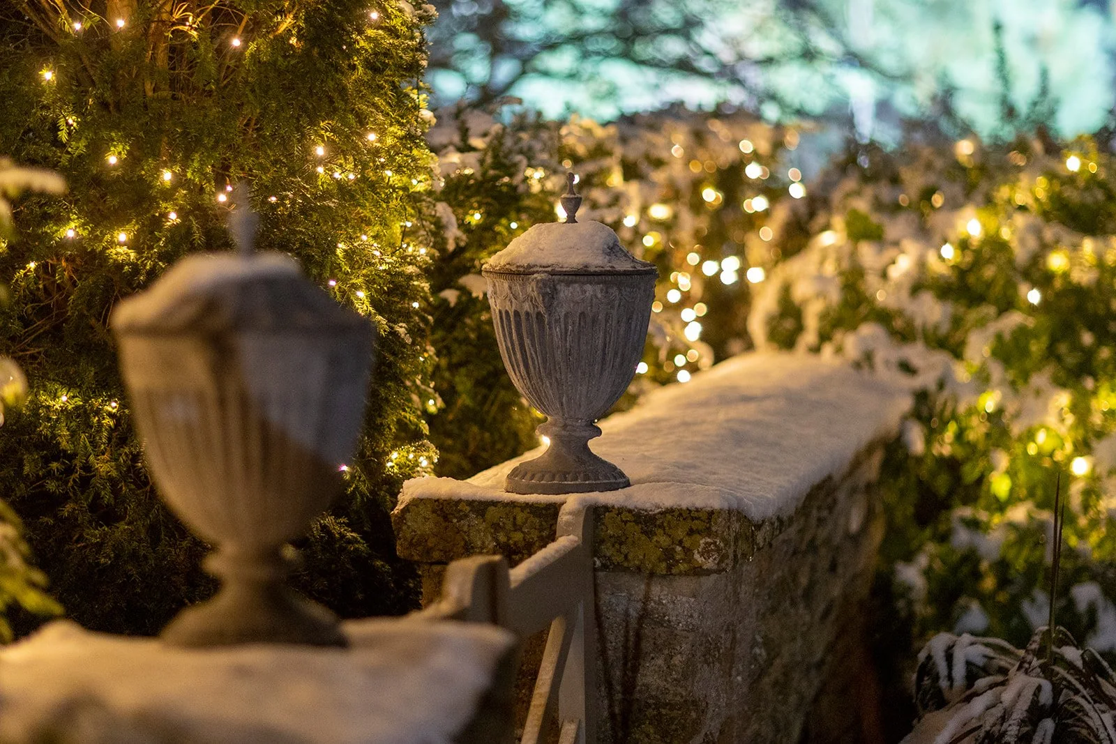 Snow-covered stone fence with decorative urns, surrounded by illuminated green bushes at night.