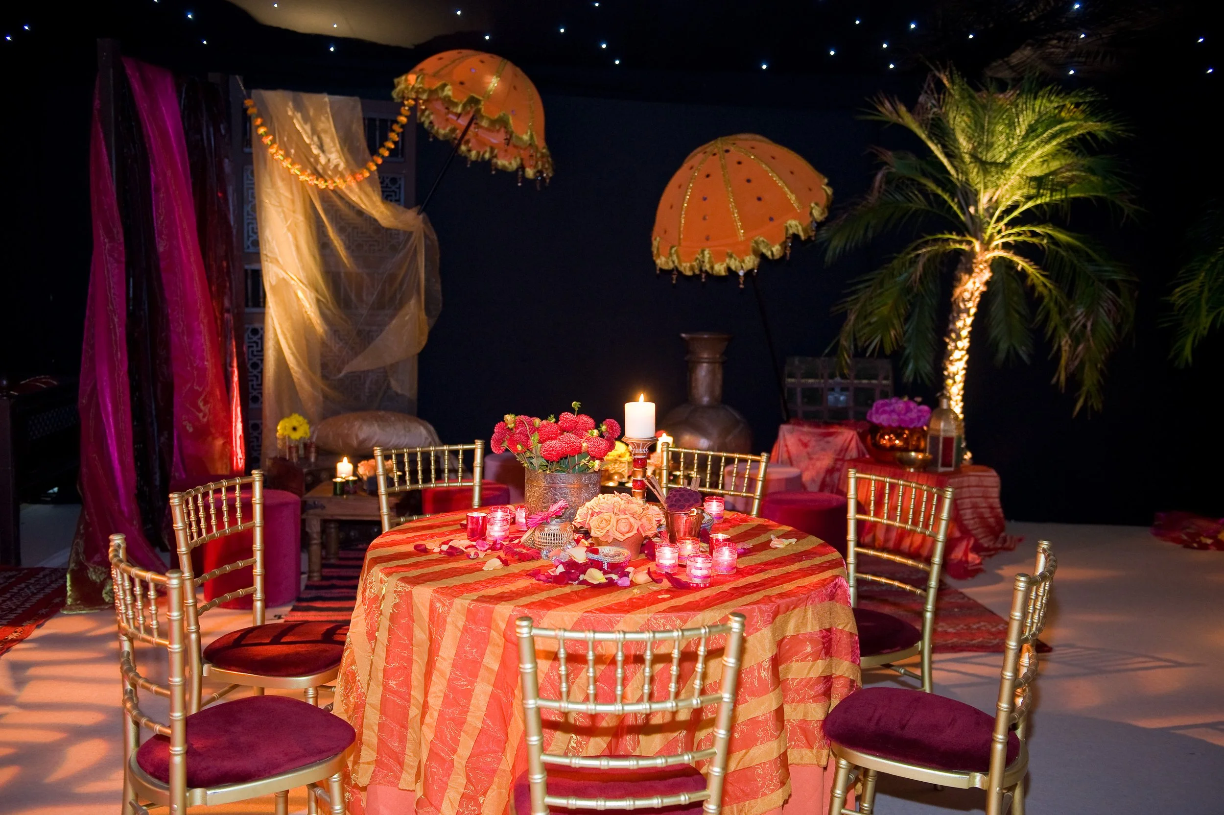 A decorated party or celebration table setup with pink flowers, candles, and rose petals. The tablecloth has a striped pattern in shades of orange and pink. Surrounding the table are gold-colored chairs with red cushions. In the background, there are