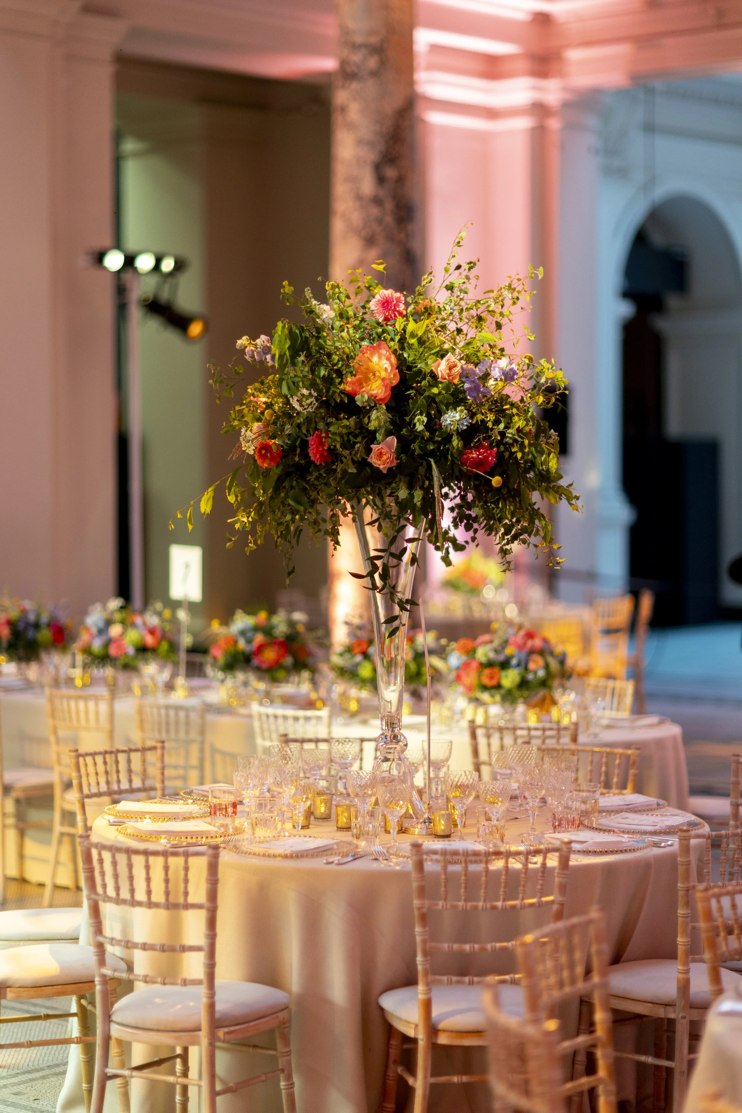 Elegant banquet setup with a tall floral centerpiece featuring pink, purple, and white flowers on a round table with glassware and Chiavari chairs, in a decorated event space.