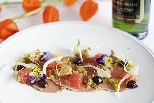 A plate of thinly sliced raw meat topping with edible flowers, small vegetables, and blackberries, garnished with microgreens, served with a bottle of green wine in the background.
